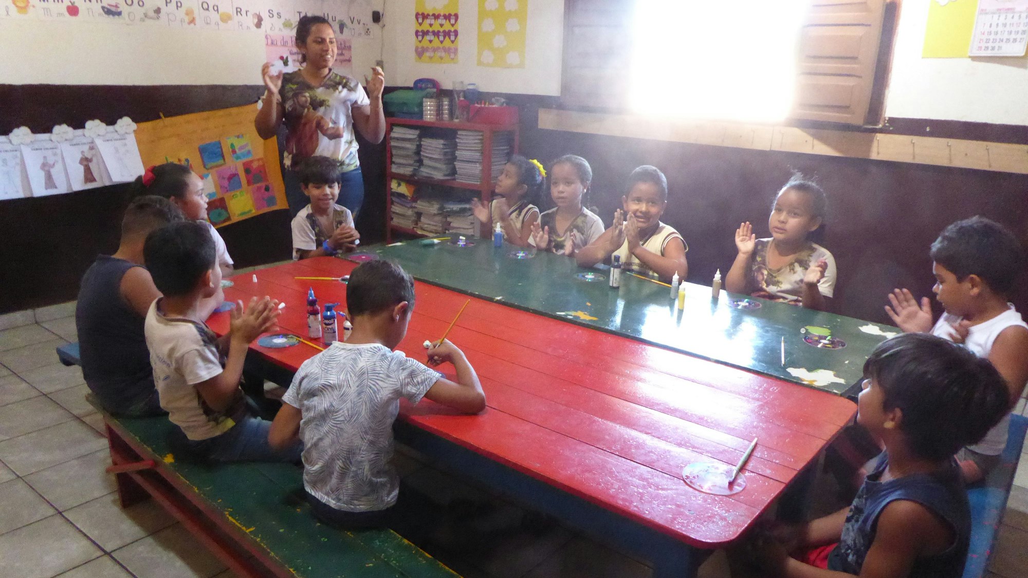 In einer Klasse sitzen zehn Schüler um einen großen Tisch. Die Lehrerin steht daneben.