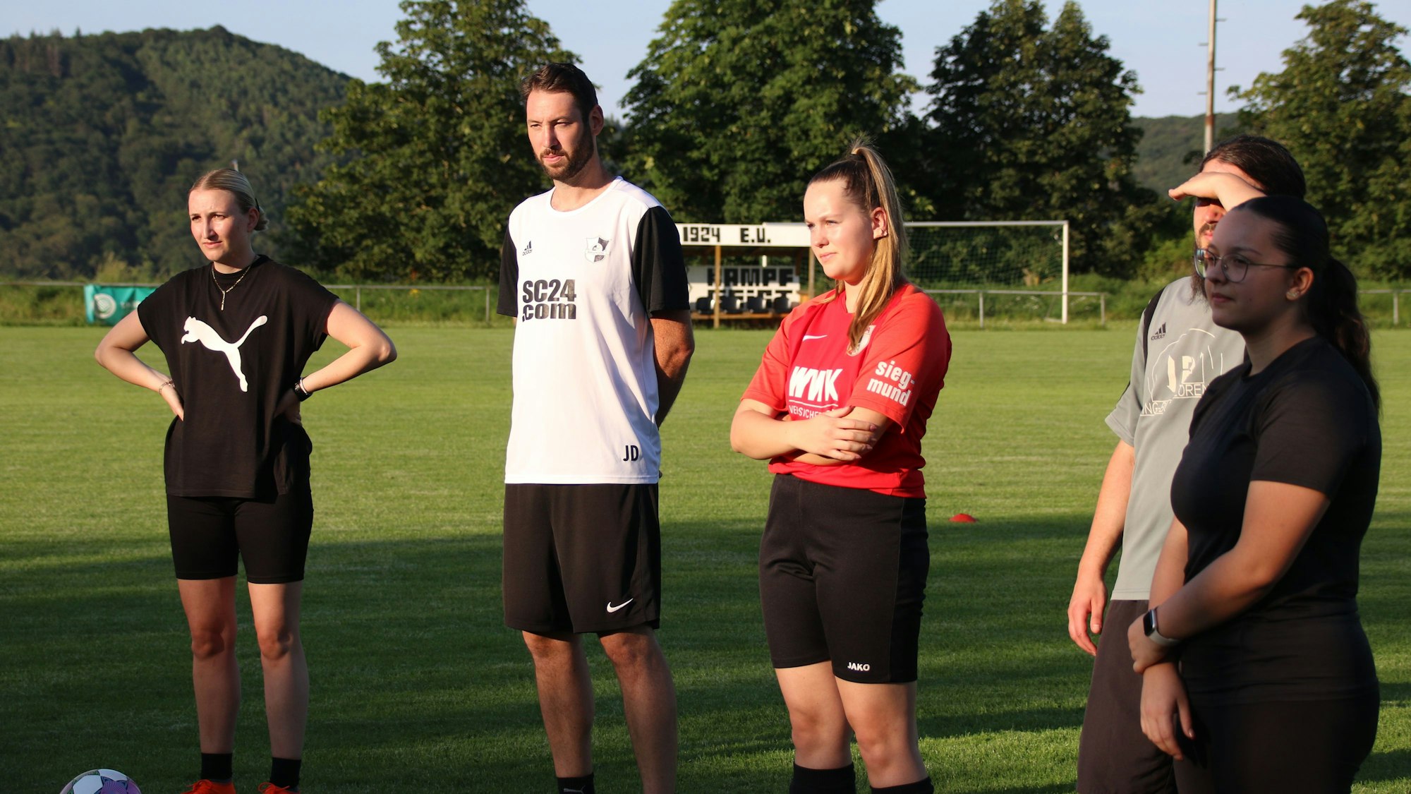 Das Bild zeigt zwei Fußballer und drei Fußballerinnen, wie sie den Worten des Trainers lauschen.