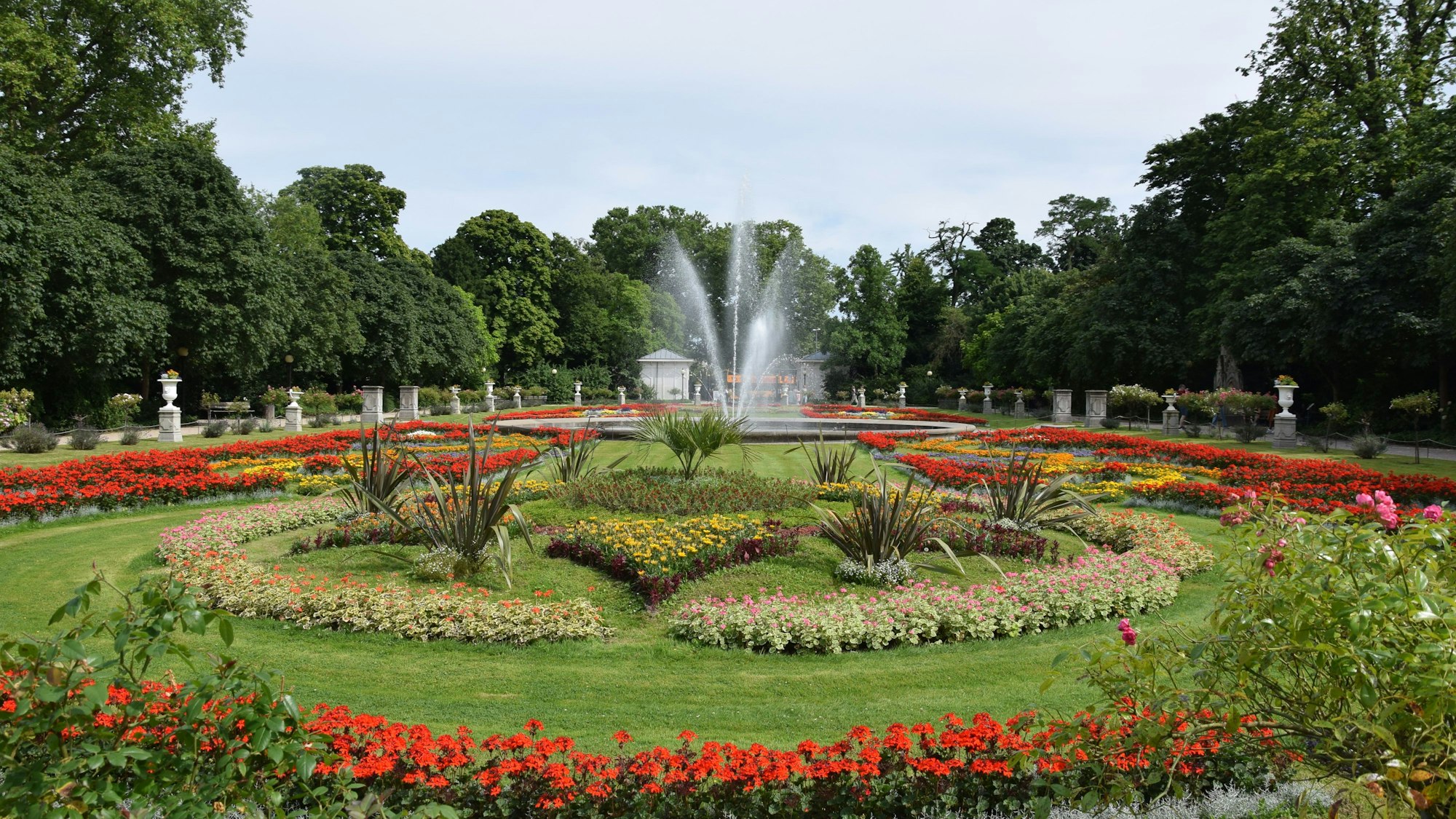 Der bunt blühende Vorgarten der Flora, mit seinen ordentlich angelegten Blumen und dem Brunnen.
