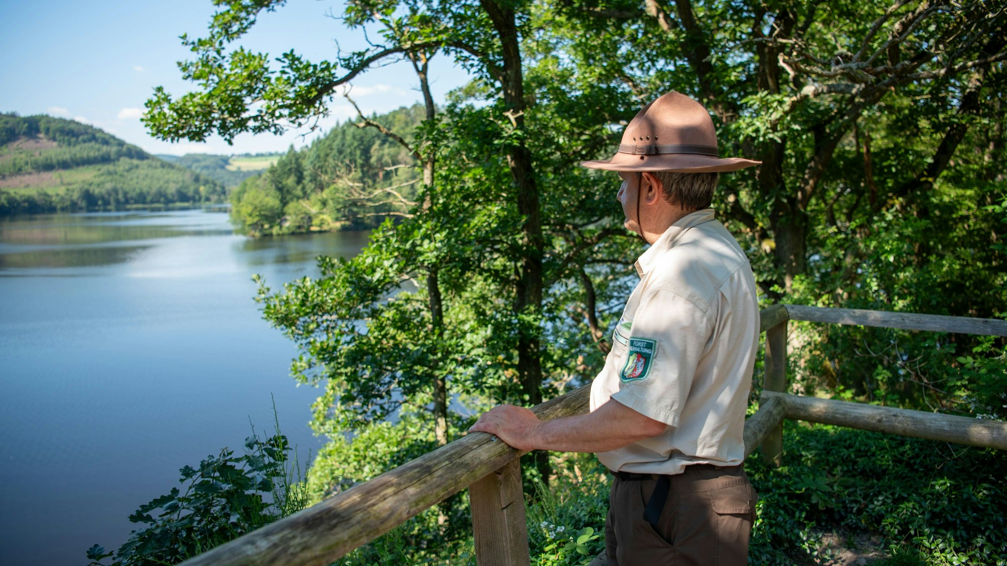 Ranger Ralf Hilgers steht in dunkelbrauner Hose, beigem Hemd und braunem Rangerhut an einem Holzgeländer und schaut auf den Rursee.