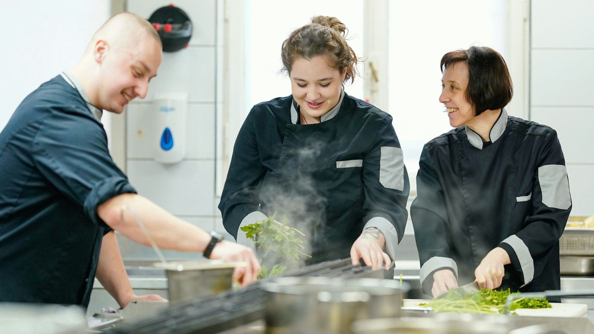 Zwei Frauen und ein Mann stehen in einer Küche und schneiden Kräuter.
