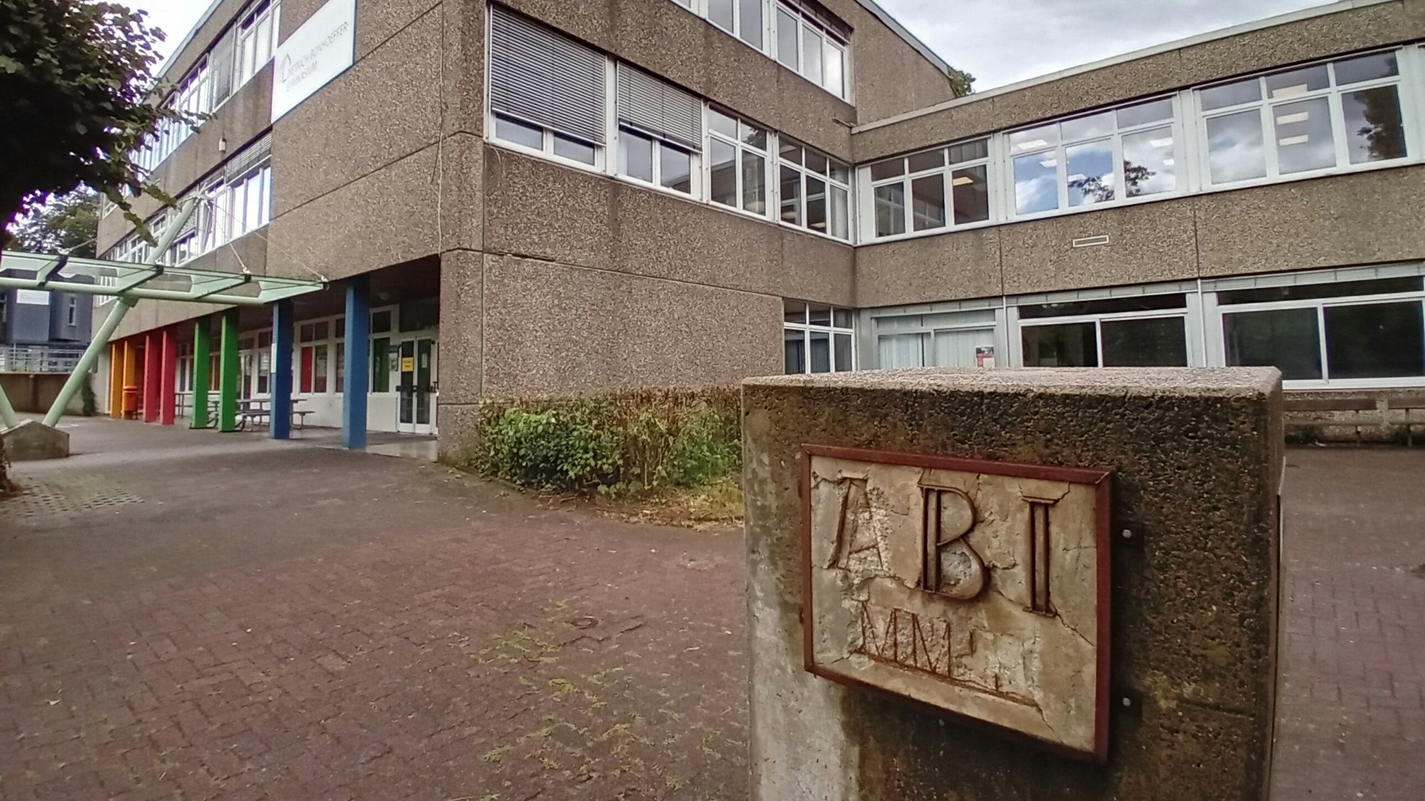 Blick auf das Schulgebäude, im Vordergrund ein Betonblock mit verwitterter Inschrift "Abi".