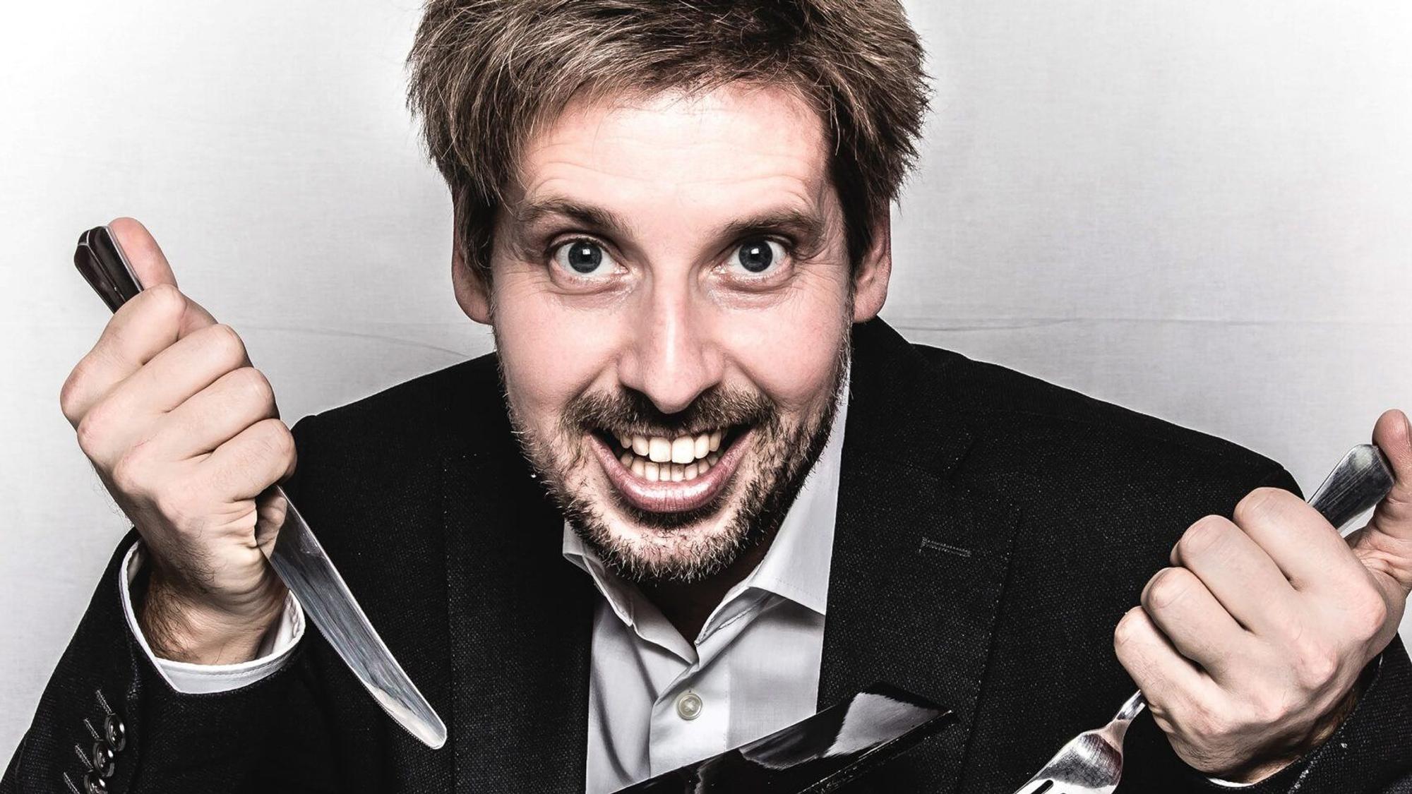 Ein Mann lacht und hält Messer und Gabel in den Händen.