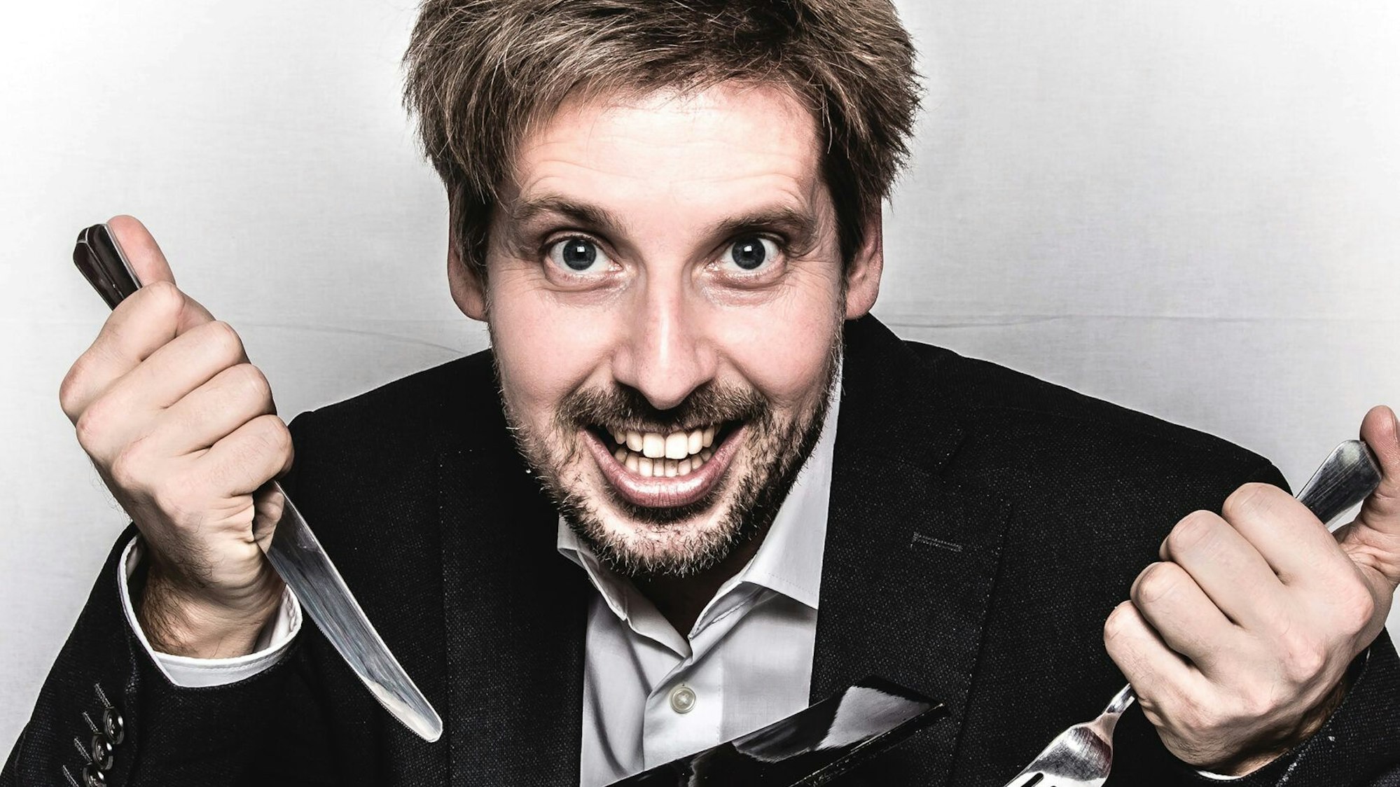 Ein Mann lacht und hält Messer und Gabel in den Händen.