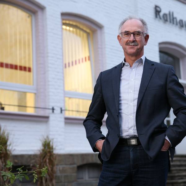Ein Mann im Anzug, mit Brille und Schnurrbart steht vor einem weißen Gebäude. Über dem Eingang steht "Rathaus".