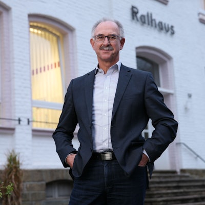 Ein Mann im Anzug, mit Brille und Schnurrbart steht vor einem weißen Gebäude. Über dem Eingang steht "Rathaus".
