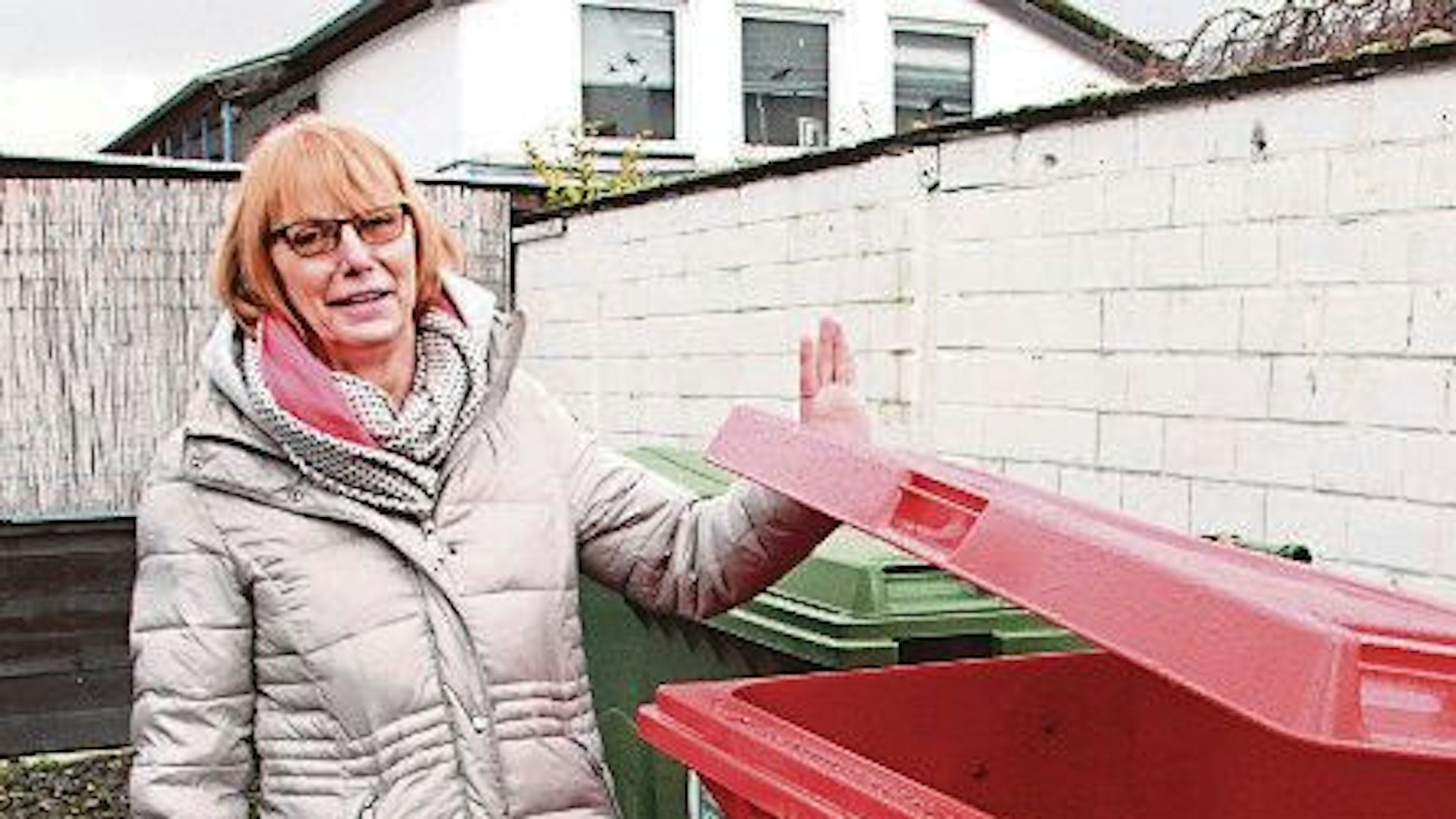 Eine Frau mit rötlichen Haaren steht neben einem roten Kunststoffcontainer. Sie trägt einen Wintermantel.