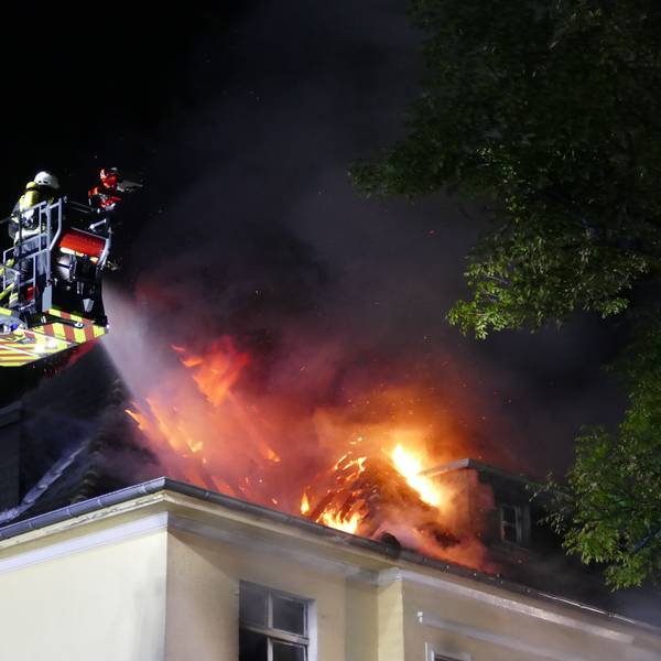Flammen schlagen aus dem Dach eines Hauses, Feuerwehrleute löschen vom Korb einer Drehleiter aus.