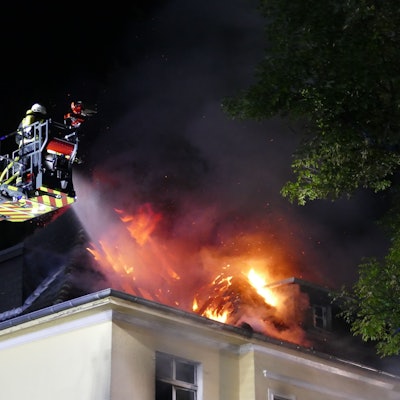 Flammen schlagen aus dem Dach eines Hauses, Feuerwehrleute löschen vom Korb einer Drehleiter aus.