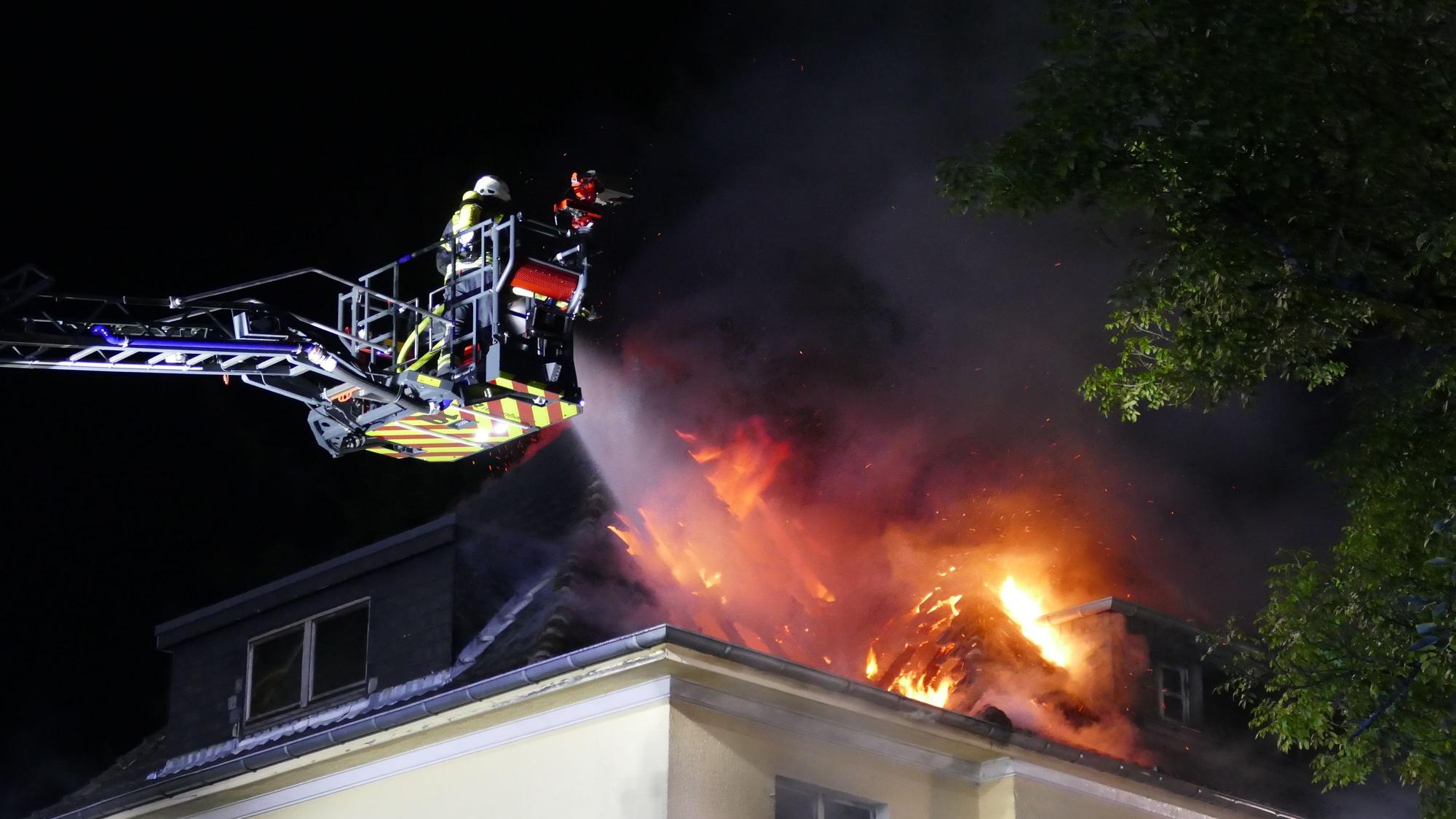 Flammen schlagen aus dem Dach eines Hauses, Feuerwehrleute löschen vom Korb einer Drehleiter aus.