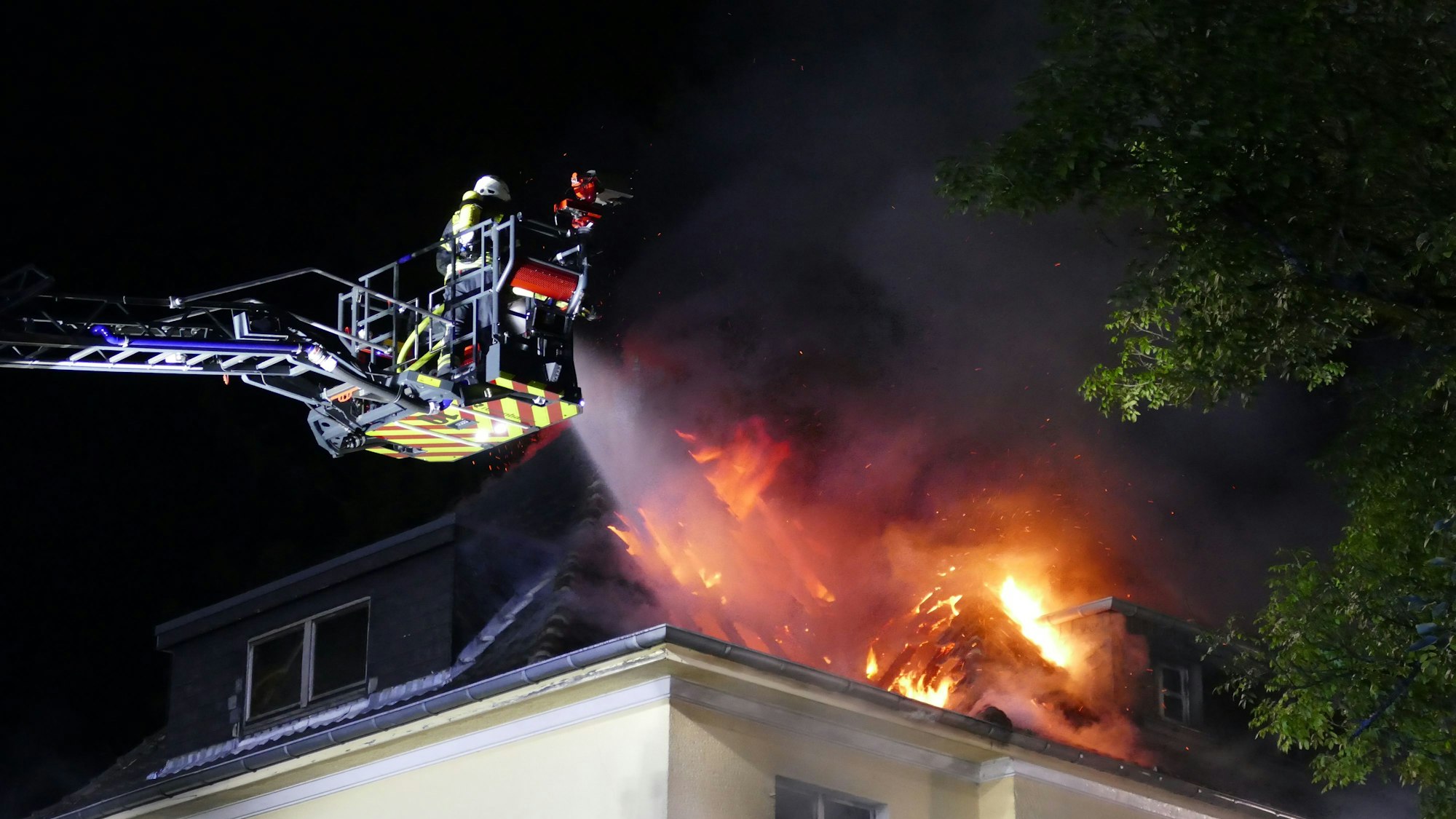 Flammen schlagen aus dem Dach eines Hauses, Feuerwehrleute löschen vom Korb einer Drehleiter aus.