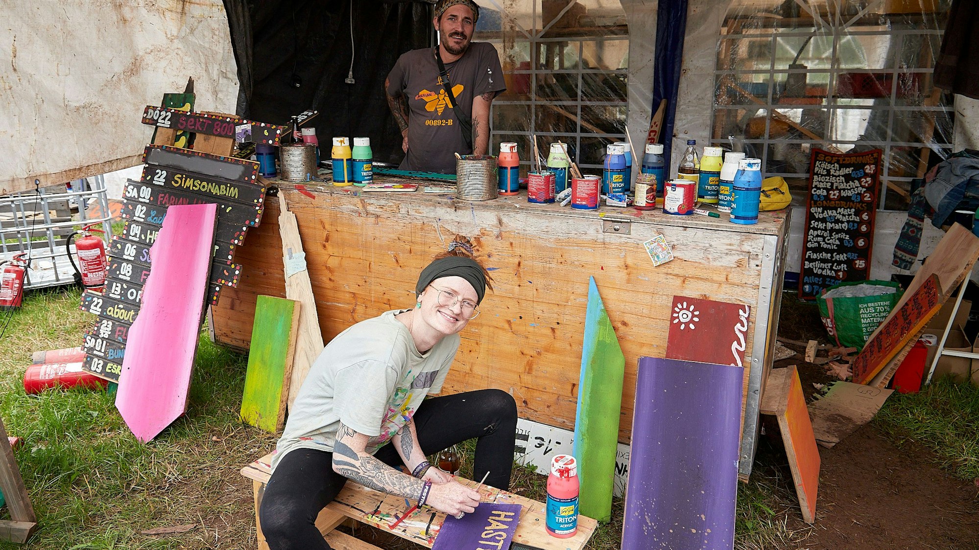 Ein Mann und eine Frau sind in einem Zelt, auf einer Theke stehen zahlreiche Farbtuben und -dosen. Sie gestalten die Hinweisschilder für das Zugvögel-Festival am Weißen Stein in Hellenthal-Udenbreth.