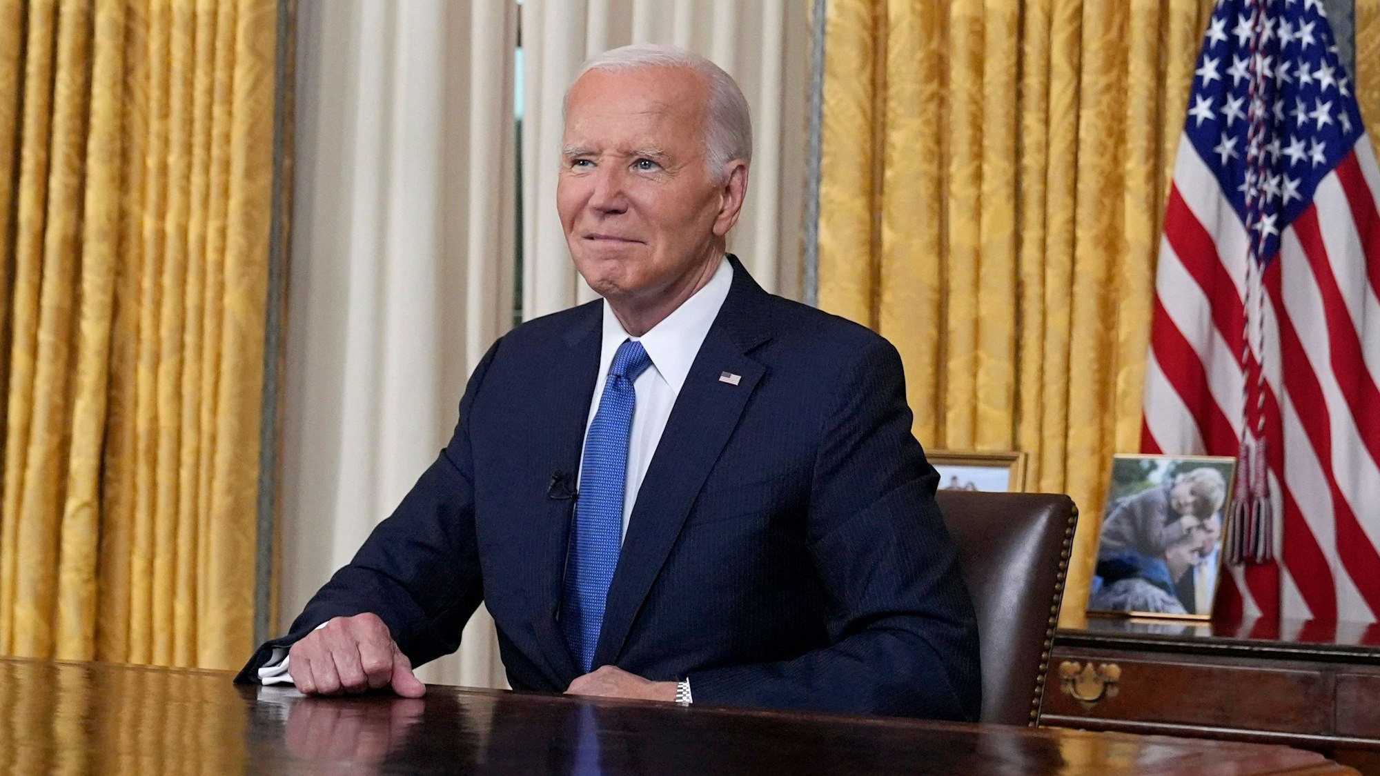 US-Präsident Joe Biden sitzt im Oval Office im Weißen Haus vor einer US-amerikanischen Flagge und blickt vor einer Rede in die Kamera.