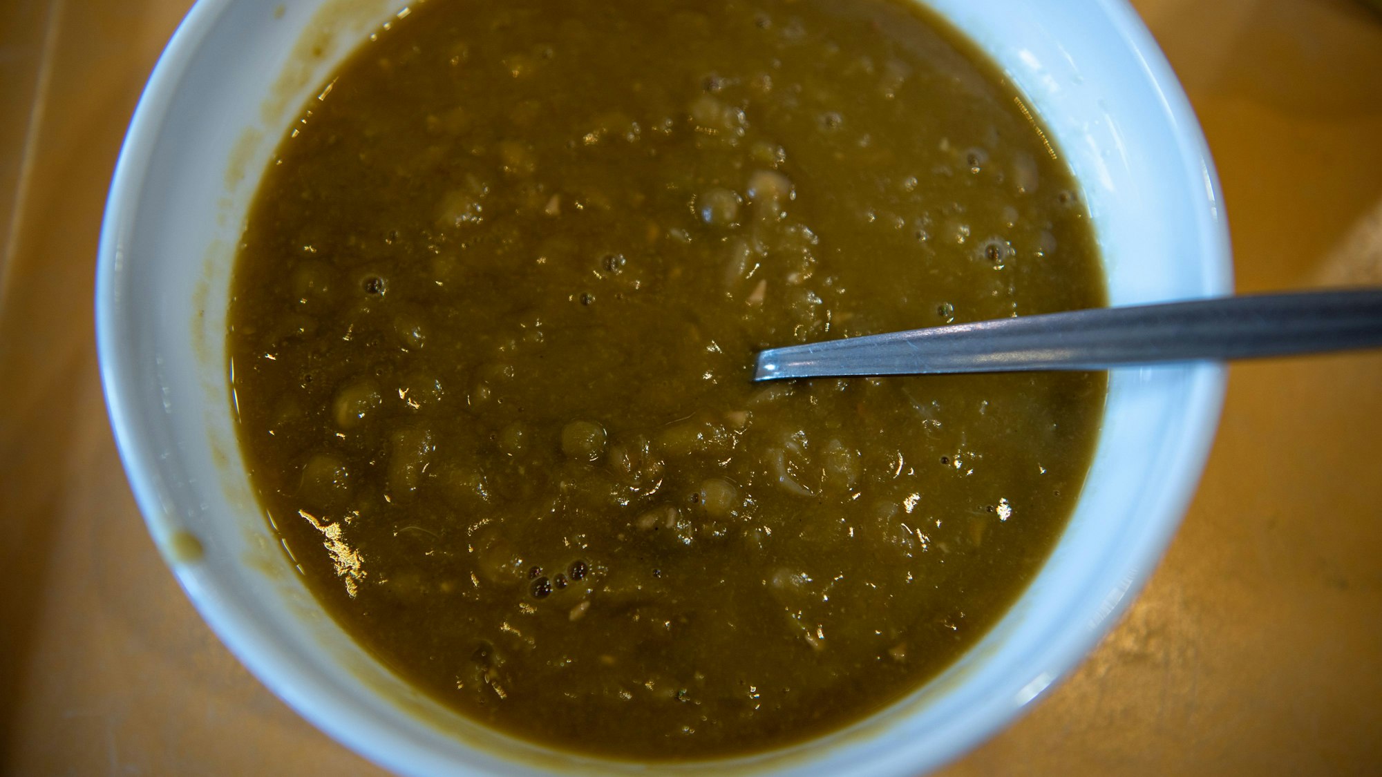 In einer weißen Schüssel befindet sich braun-grüne Erbsensuppe. In der Schüssel steckt ein Löffel.