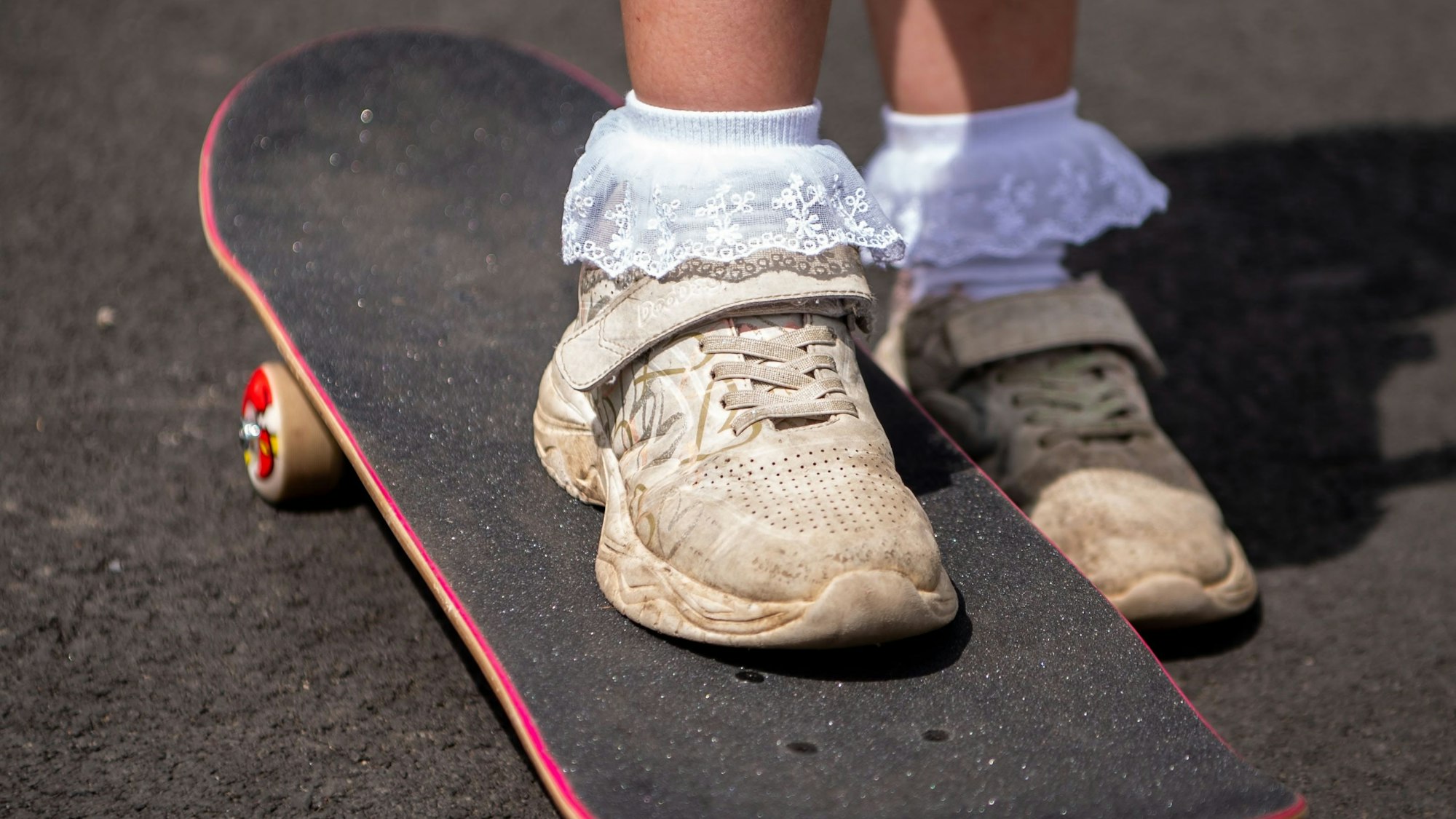 Das Bild zeigt die Füße eines Mädchens. Es trägt Rüschensocken. Das rechte Bein steht auf einem Skateboard.