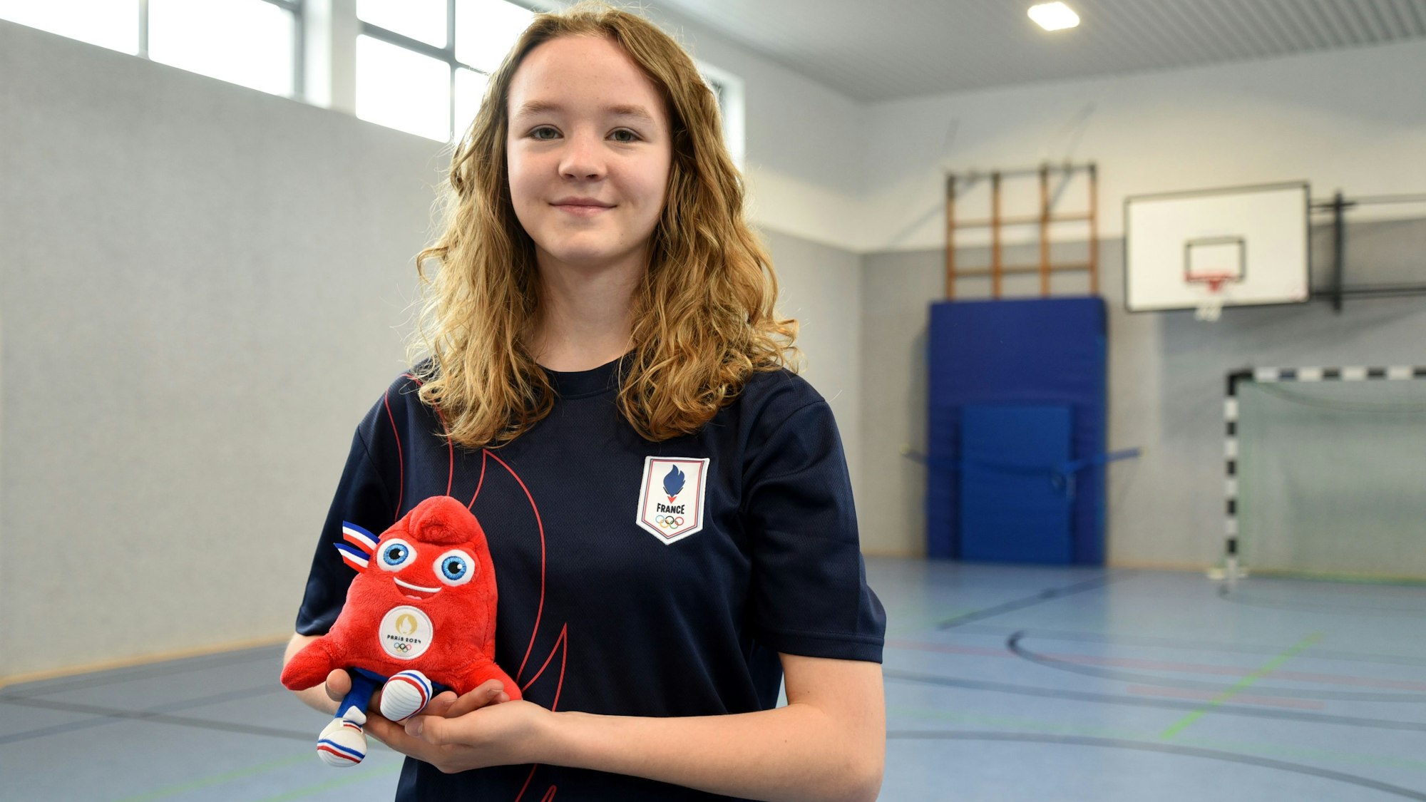 Lina Friedrichs steht in der Sporthalle des TuS Elsenroth und hat das Olympia-Maskottchen in der Hand.