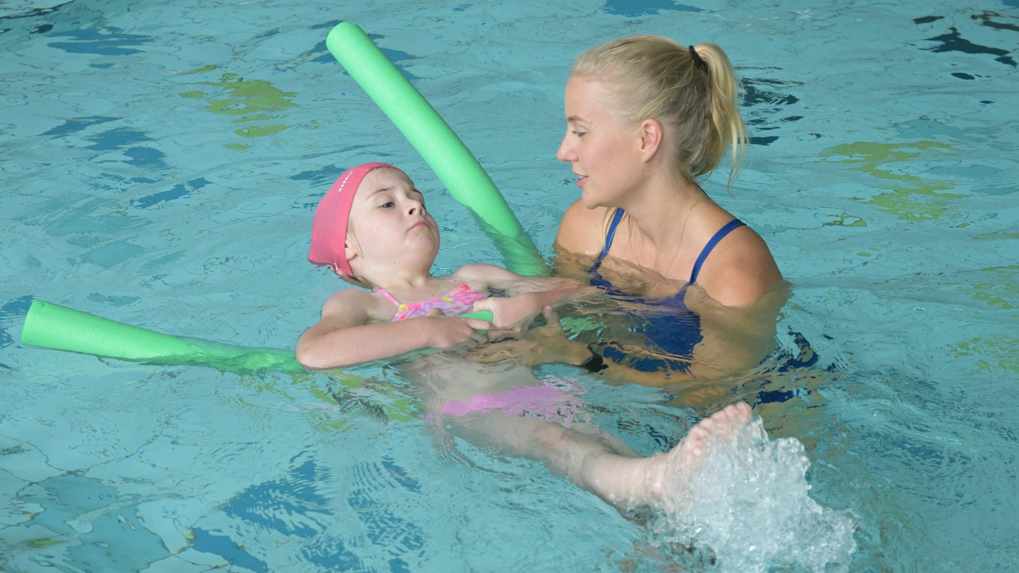 Anna liegt auf dem Rücken im Wasser und wird durch ihre Lehrerin und eine Schwimmnudel unterstützt.