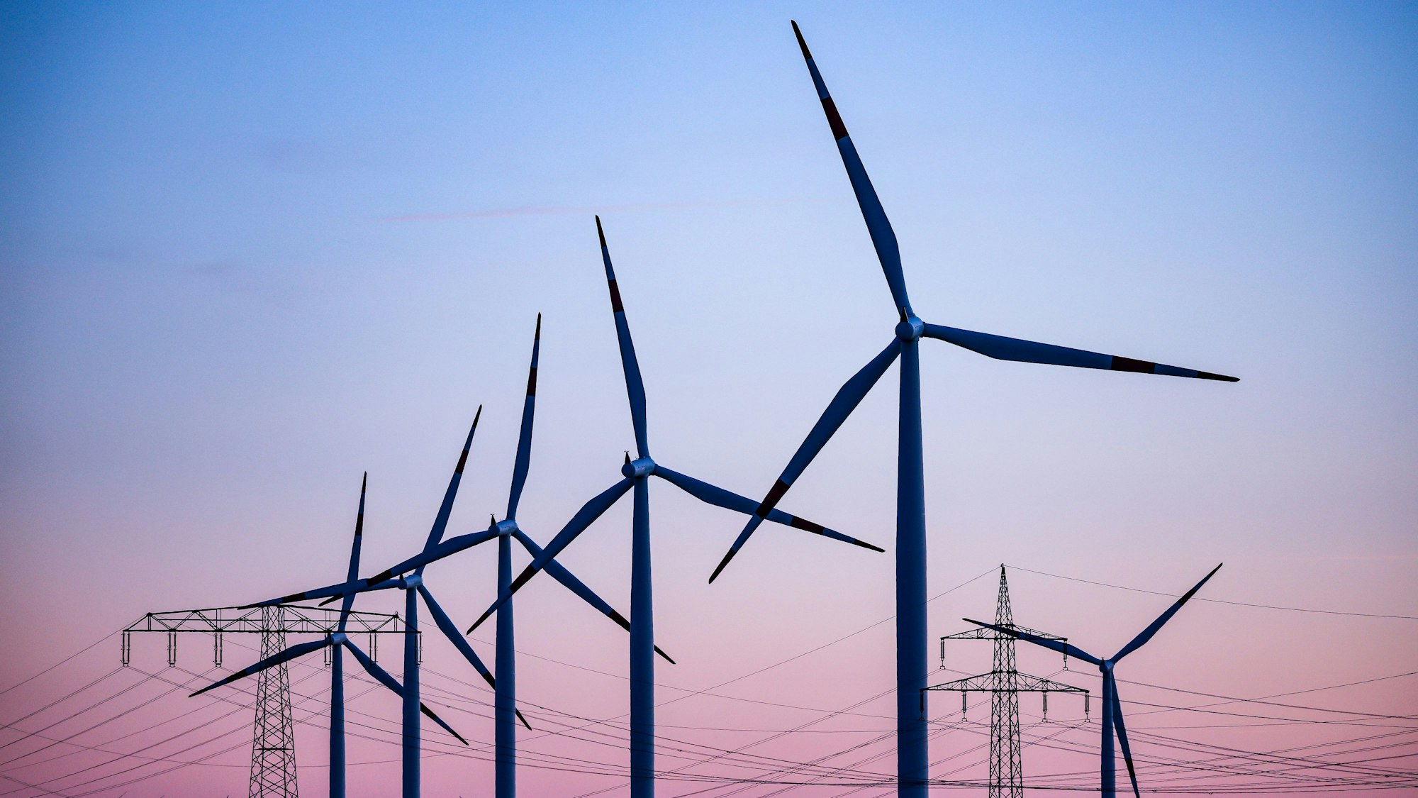 Windkraftanlagen und Hochspannungsleitungen sind vor dem Abendhimmel zu sehen. Zuletzt sorgte eine „Dunkelflaute“ für hohe Preise am Strommarkt. (Symbolbild)