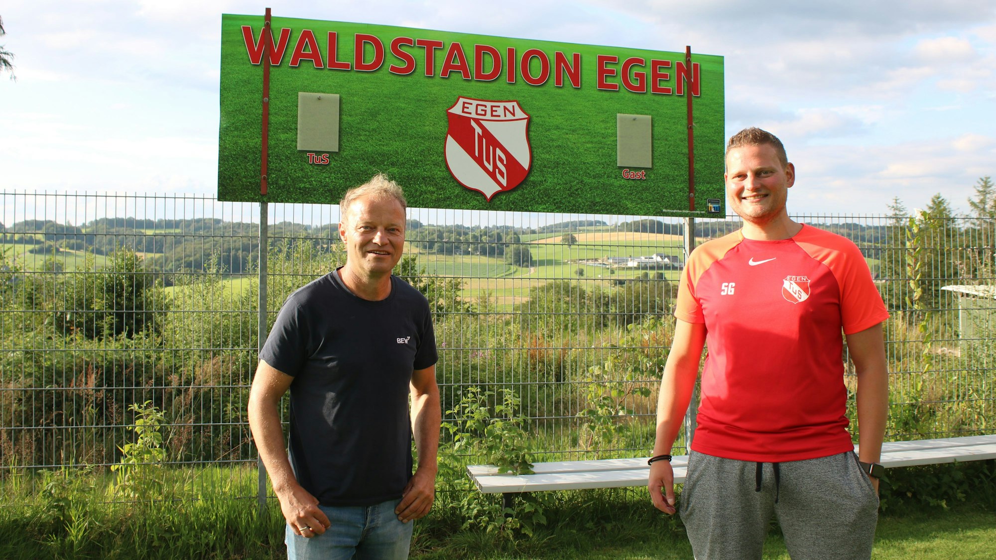 Zwei Männer stehen an einem Fußballplatz nebeneinander.
