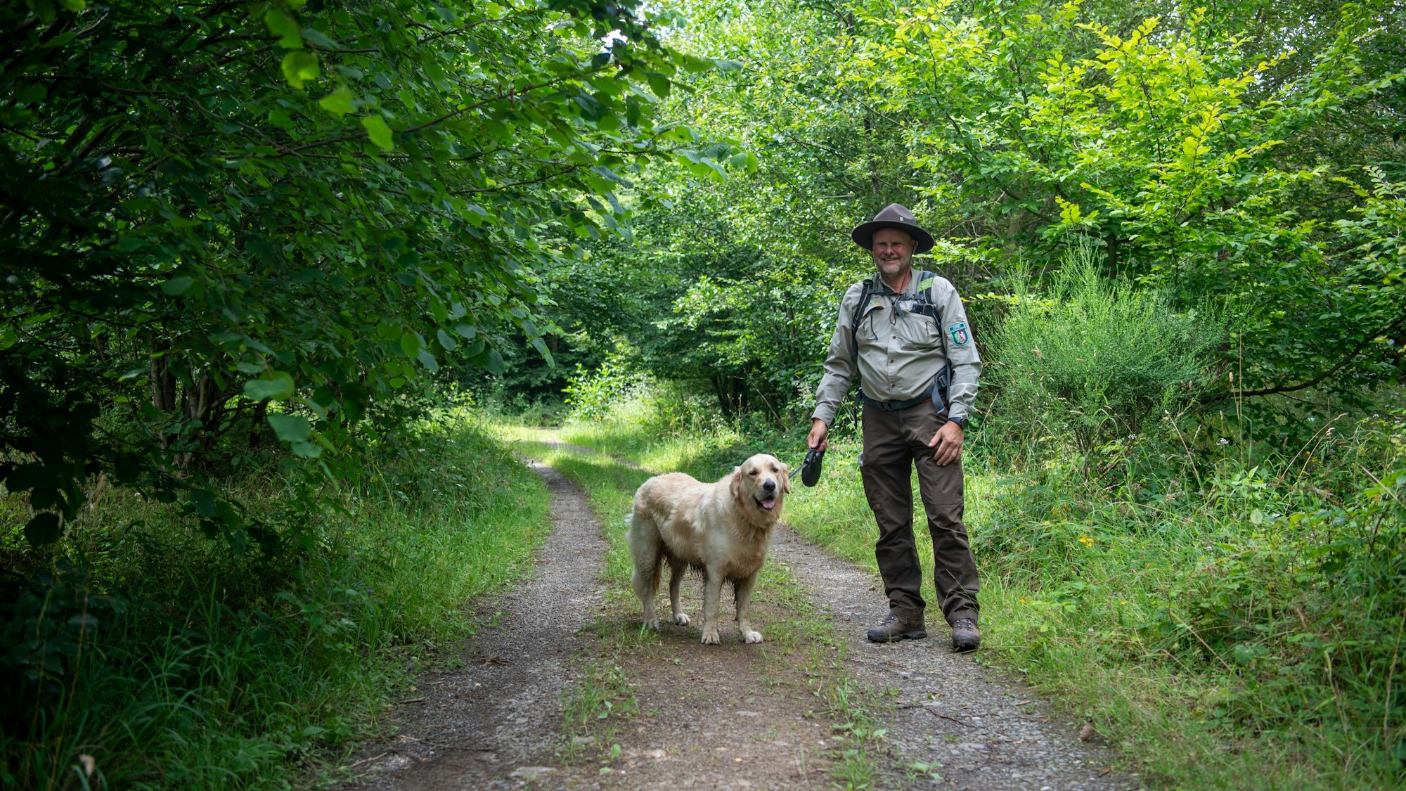 Rager Rolf Jacobs steht in brauner Wanderhose, beigem Hemd und braunem Rangerhut rechts neben seinem Hund auf einem breiten Wanderweg zwischen jungen Bäumen.