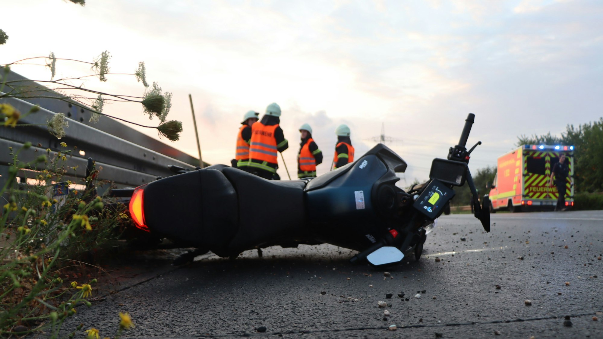 Ein Roller liegt auf der Seite auf der Straße. Im Hintergrund besprechen sich Einsatzkräfte und ein Rettungswagen steht daneben auf der Straße.