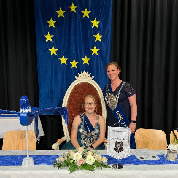 Zwei Frauen, eine sitzt, die andere steht an ihrer Seite, hinter ihnen hängt die Europafahne.