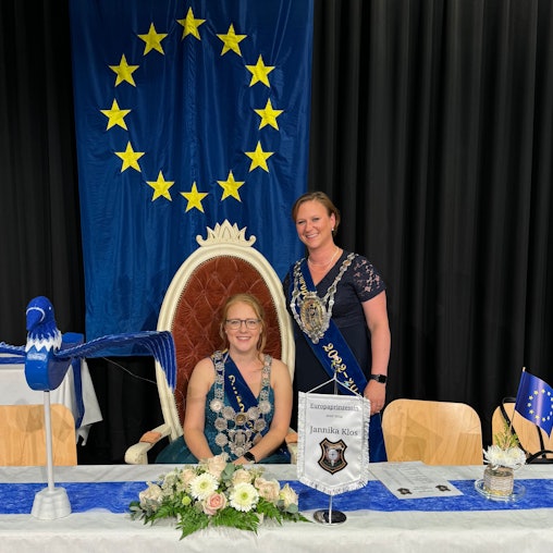 Zwei Frauen, eine sitzt, die andere steht an ihrer Seite, hinter ihnen hängt die Europafahne.