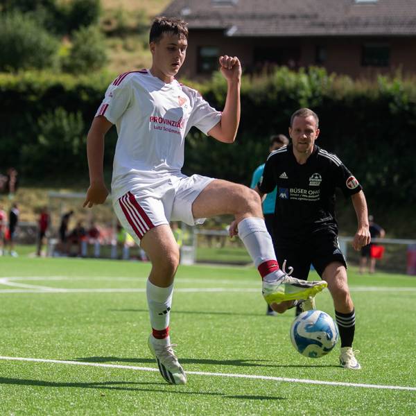Ein junger Mann in einem weißen Fußballtrikot schaut auf den Fußball, der über den Rasen des Fußballplatzes in seine Richtung fliegt.