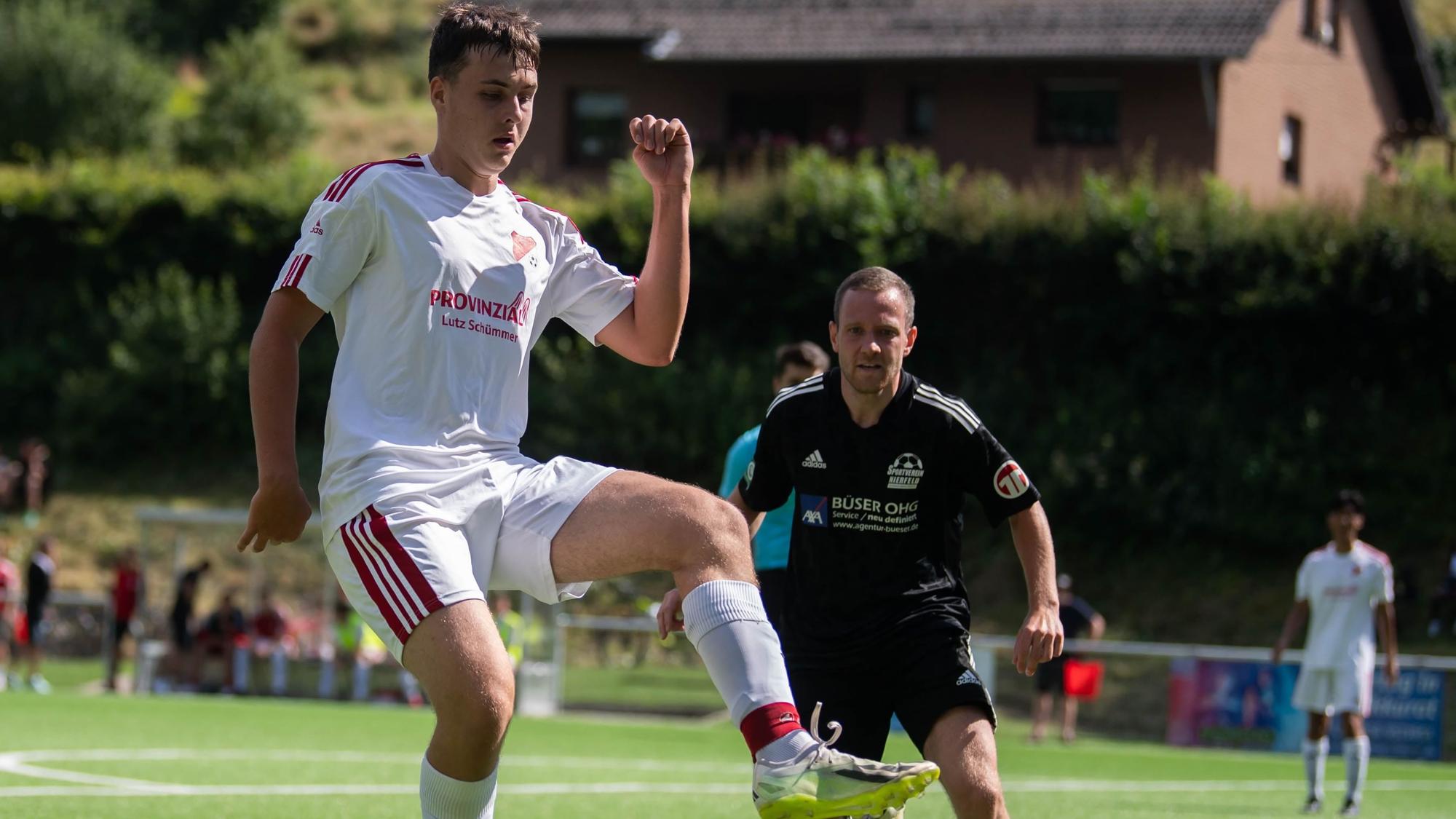 Ein junger Mann in einem weißen Fußballtrikot schaut auf den Fußball, der über den Rasen des Fußballplatzes in seine Richtung fliegt.