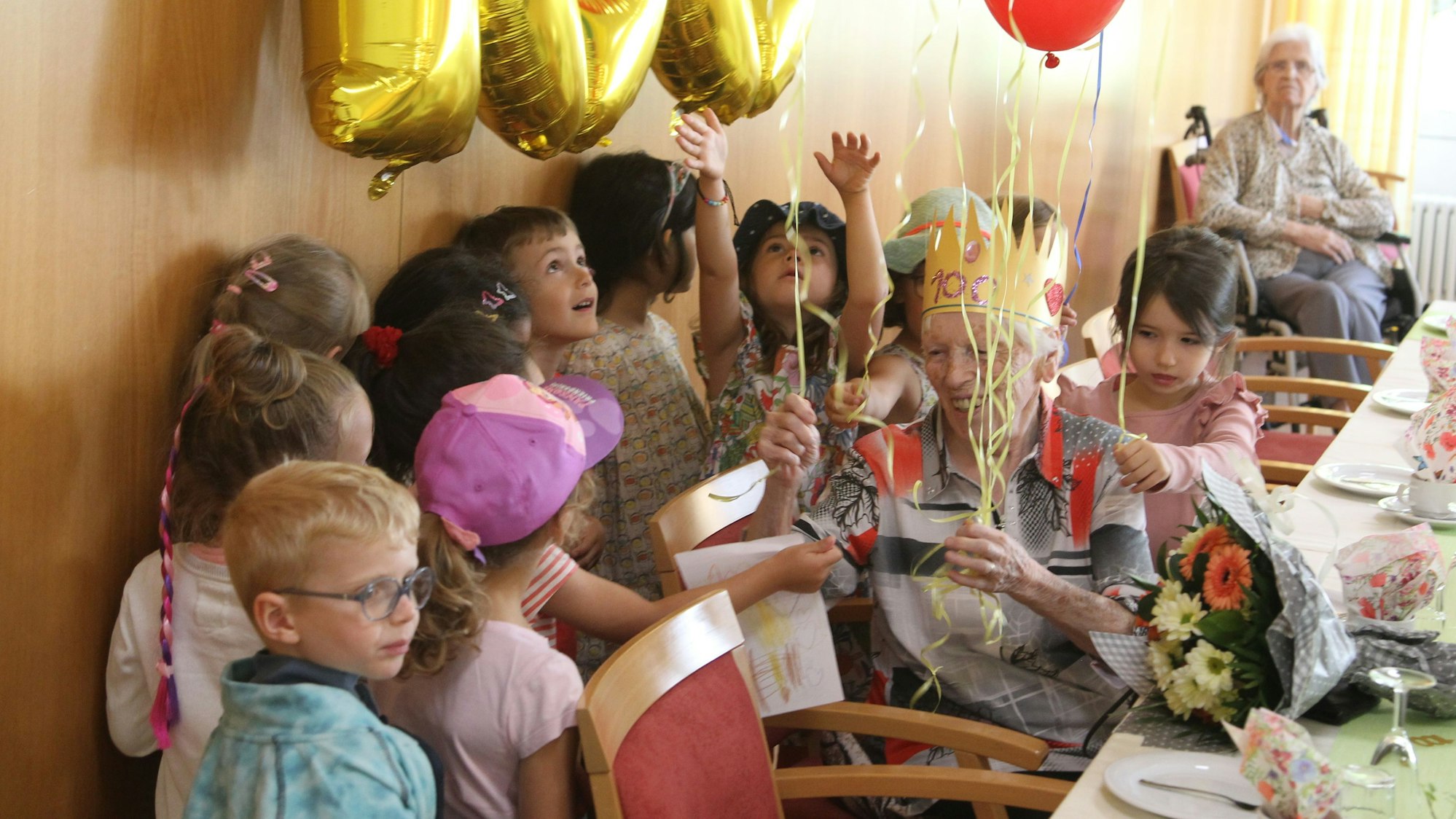 Kinder mit Luftballons in der Hand gratulieren einer alten Dame, die mit einer Krone auf dem Kopf an einem Tisch sitzt.