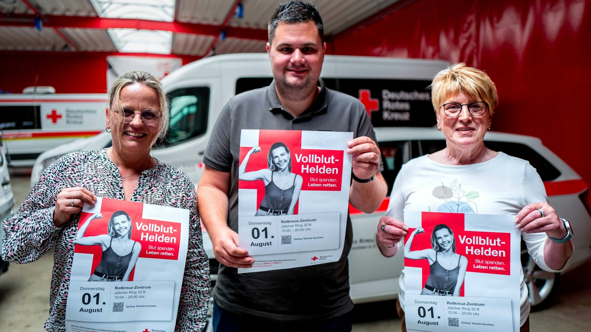 Die DRK-Mitarbeiter Stefanie Berg, Daniel Pöthmann und Edeltraud Engelen halten Plakate in der Hand, mit denen um Blutspender geworben wird. Im Hintergrund sind Einsatzfahrzeuge des DRK zu sehen.