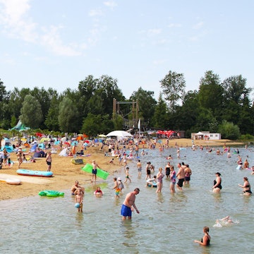 Viele Menschen in Badekleidung stehen im Wasser oder liegen auf Sandstrand, im Hintergrund sind Bäume zu sehen.