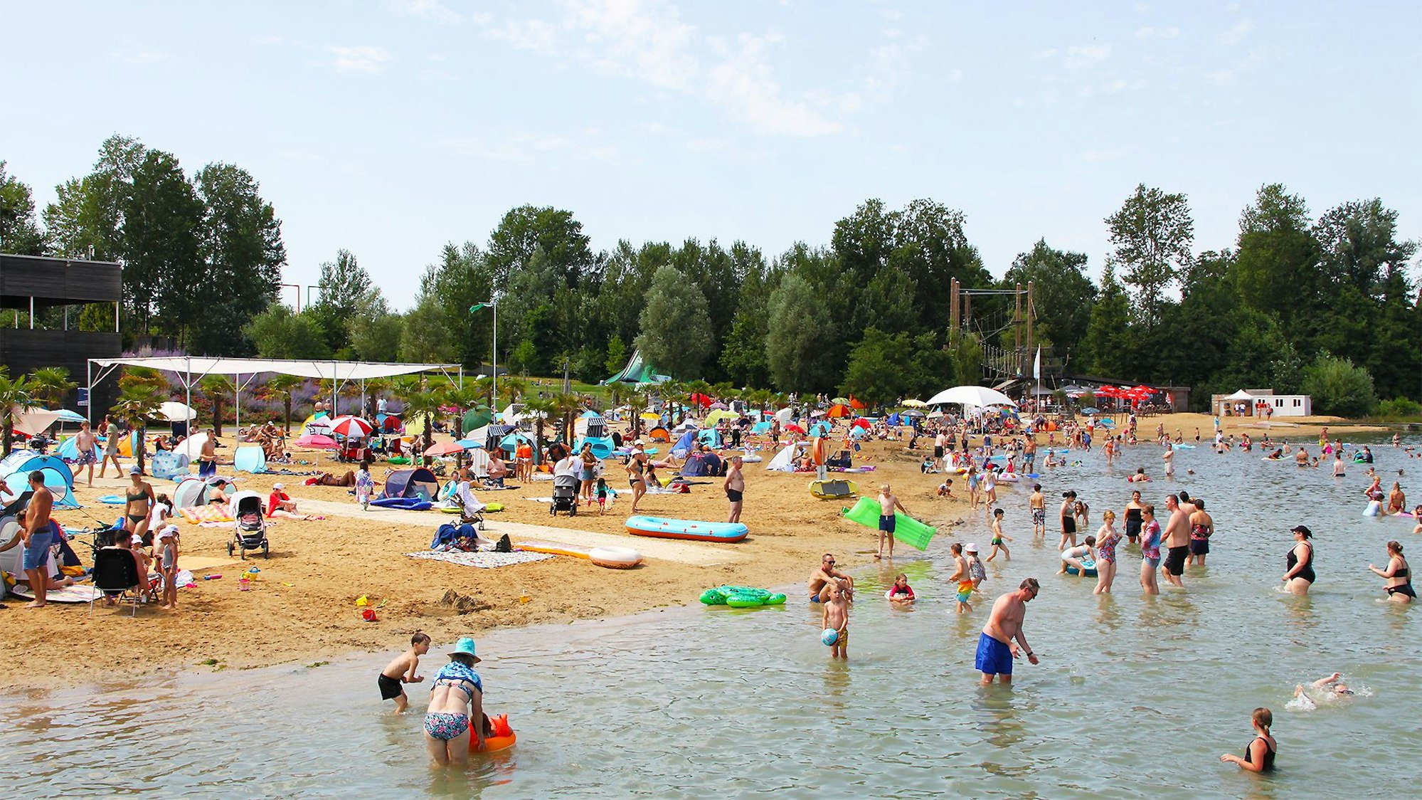 Viele Menschen in Badekleidung stehen im Wasser oder liegen auf Sandstrand, im Hintergrund sind Bäume zu sehen.