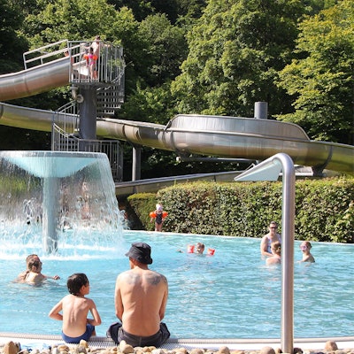 Badegäste tummeln sich an und in einem Nichtschwimmerbecken, im Hintergrund ist eine Rutsche zu sehen.