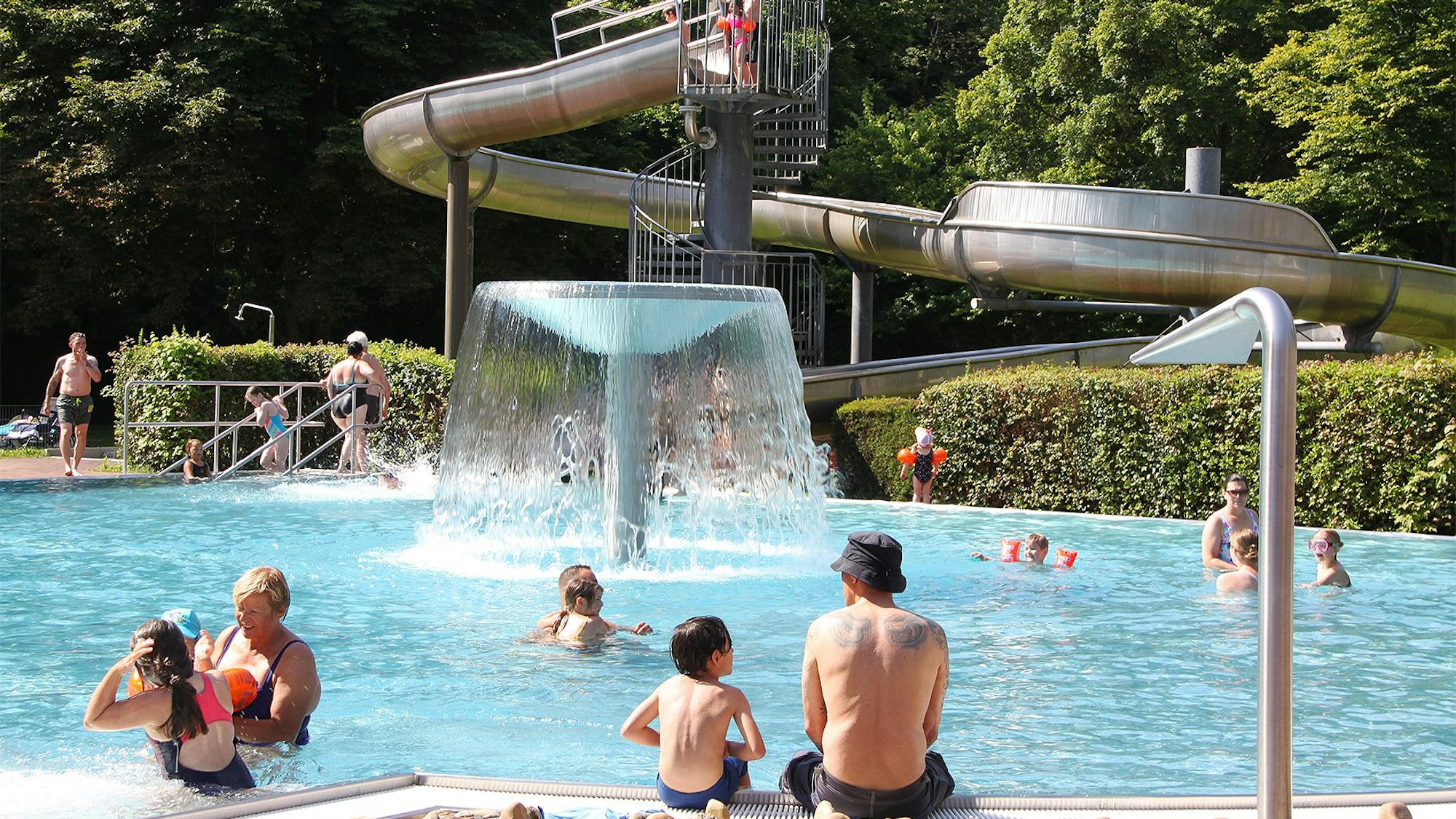 Badegäste tummeln sich an und in einem Nichtschwimmerbecken, im Hintergrund ist eine Rutsche zu sehen.
