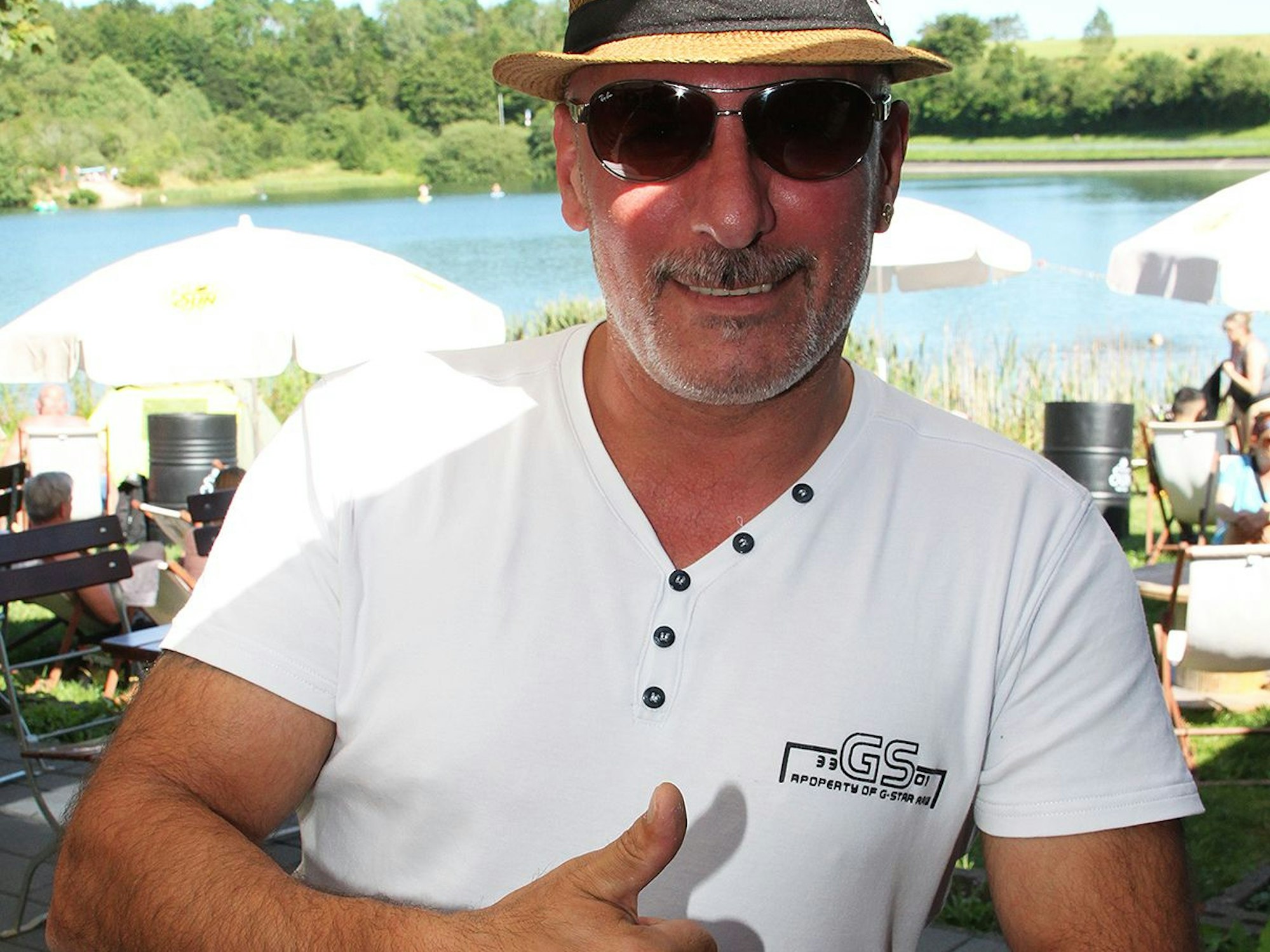 Gastronom Setin Ergogan steht an seiner See-Bar am Freilinger See. Er trägt einen Strohhut und reckt einen Daumen in die Höhe.