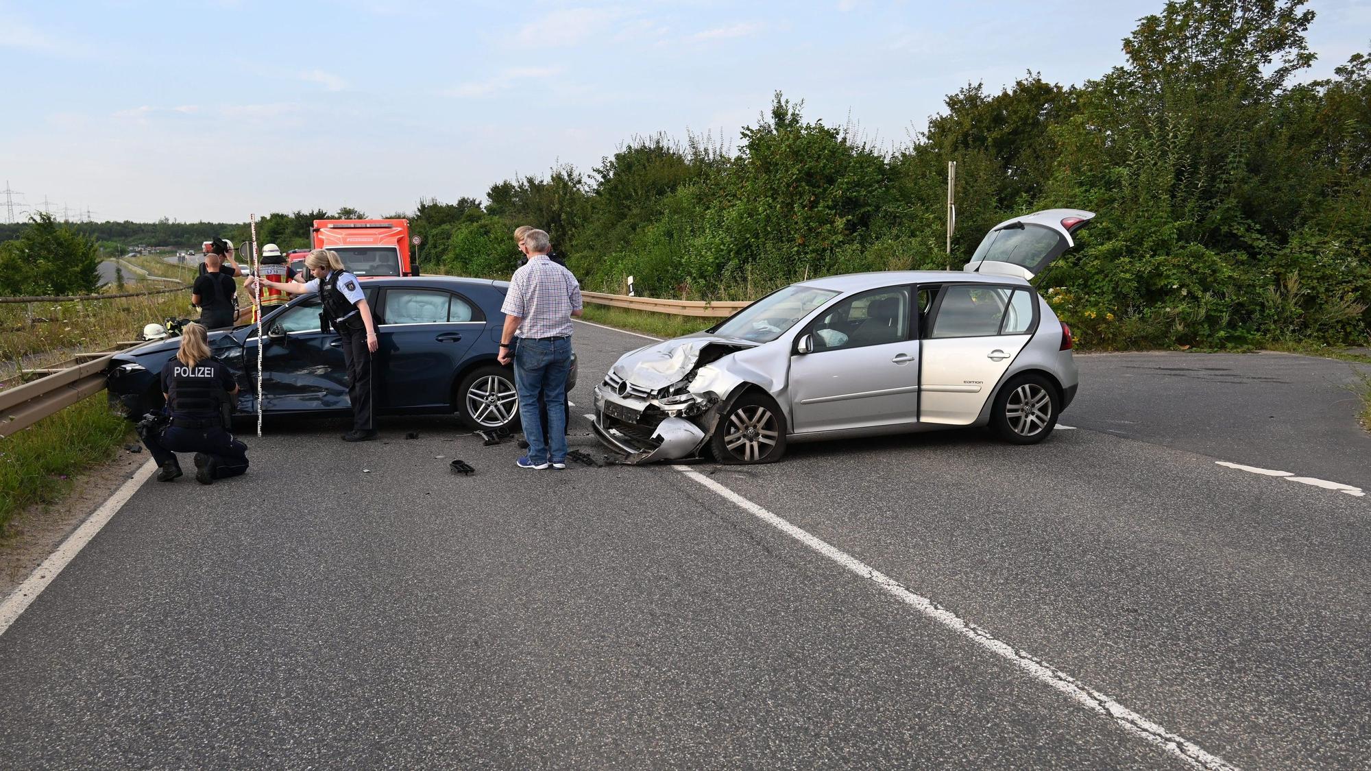 Zwei beschädigte Fahrzeuge stehen quer auf einer Straße. Auf der Fahrbahn stehen Personen, unter anderem Einsatzkräfte der Polizei. Im Hintergrund ein Rettungsfahrzeug.