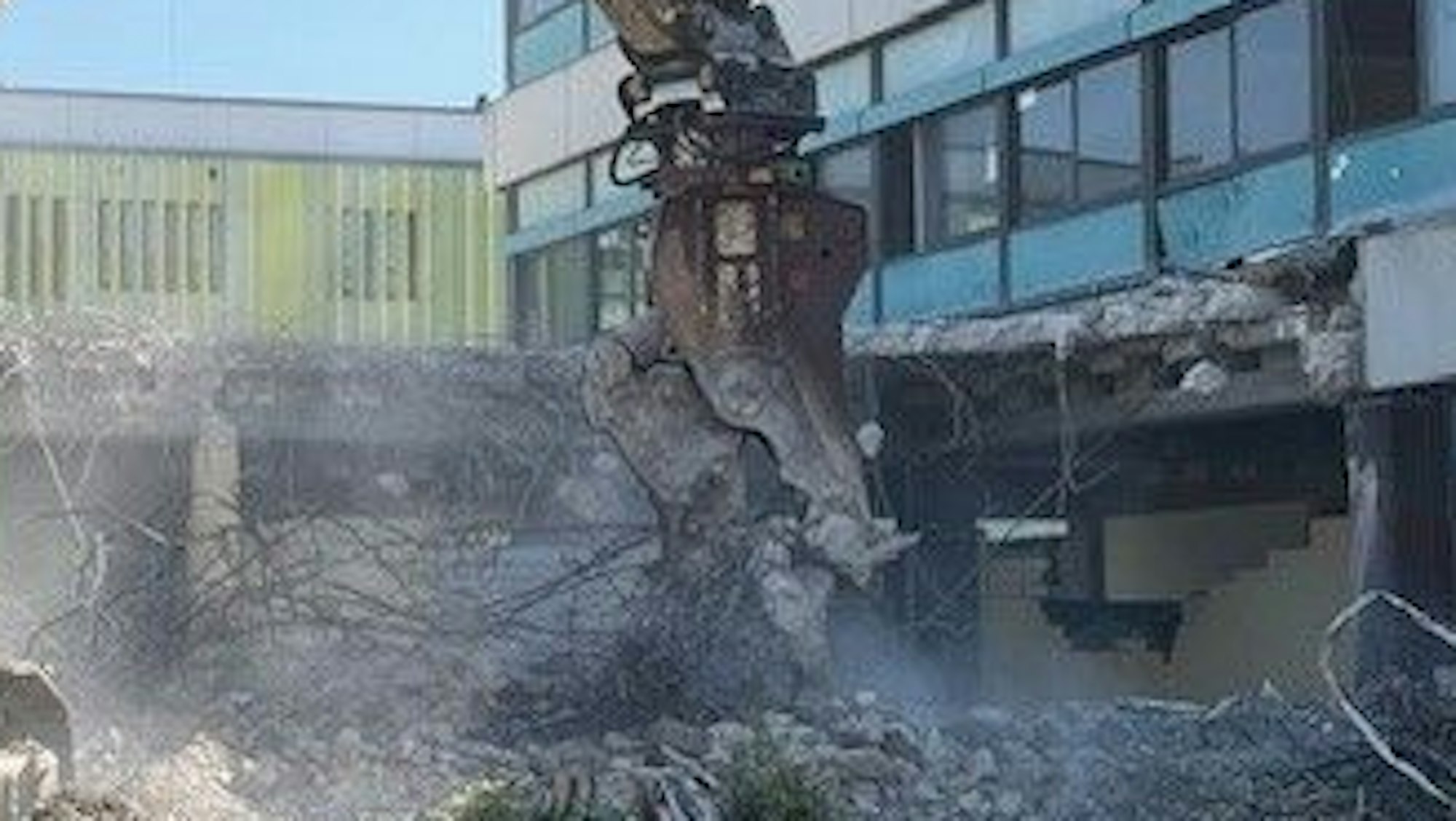 Eine Baggerzange bricht Stücke aus einem 70er-Jahre-Gebäude.