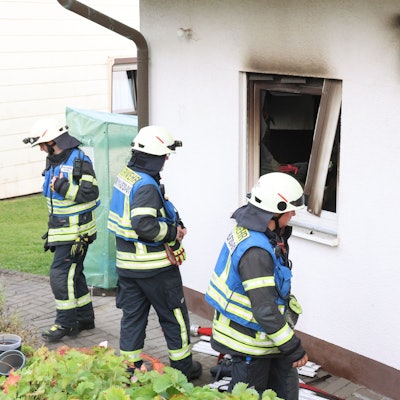 Drei Feuerwehrleute stehen vor einem Haus, in dem es zuvor gebrannt hat.