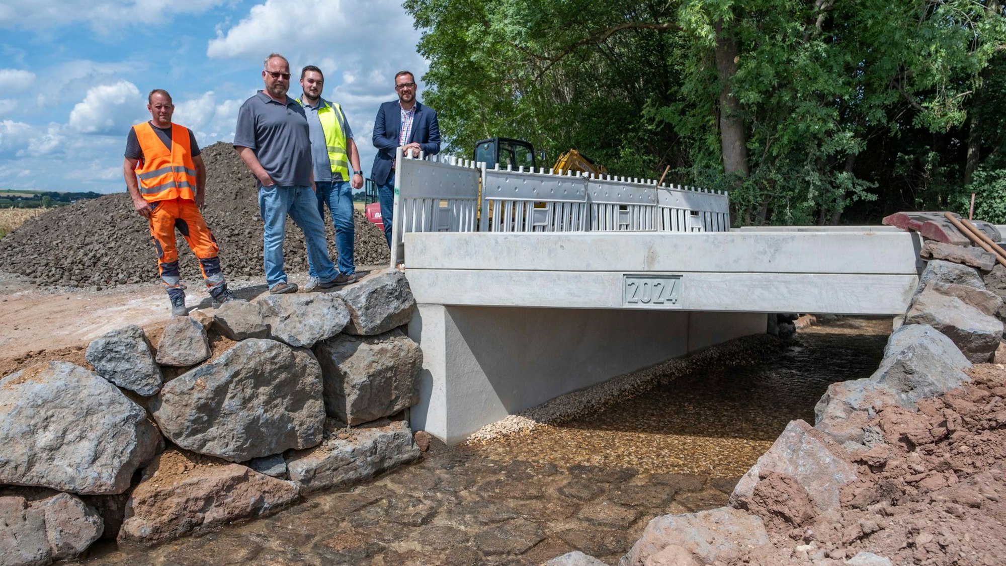Thomas Hambach (v.r.), Tiefbausachbearbeiter Marvin Perschke, Teamleiter Andreas König und Polier Jens Plinius stehen an einer Betonbrücke mit der Jahreszahl 2024, die über einen kleinen Bach führt.