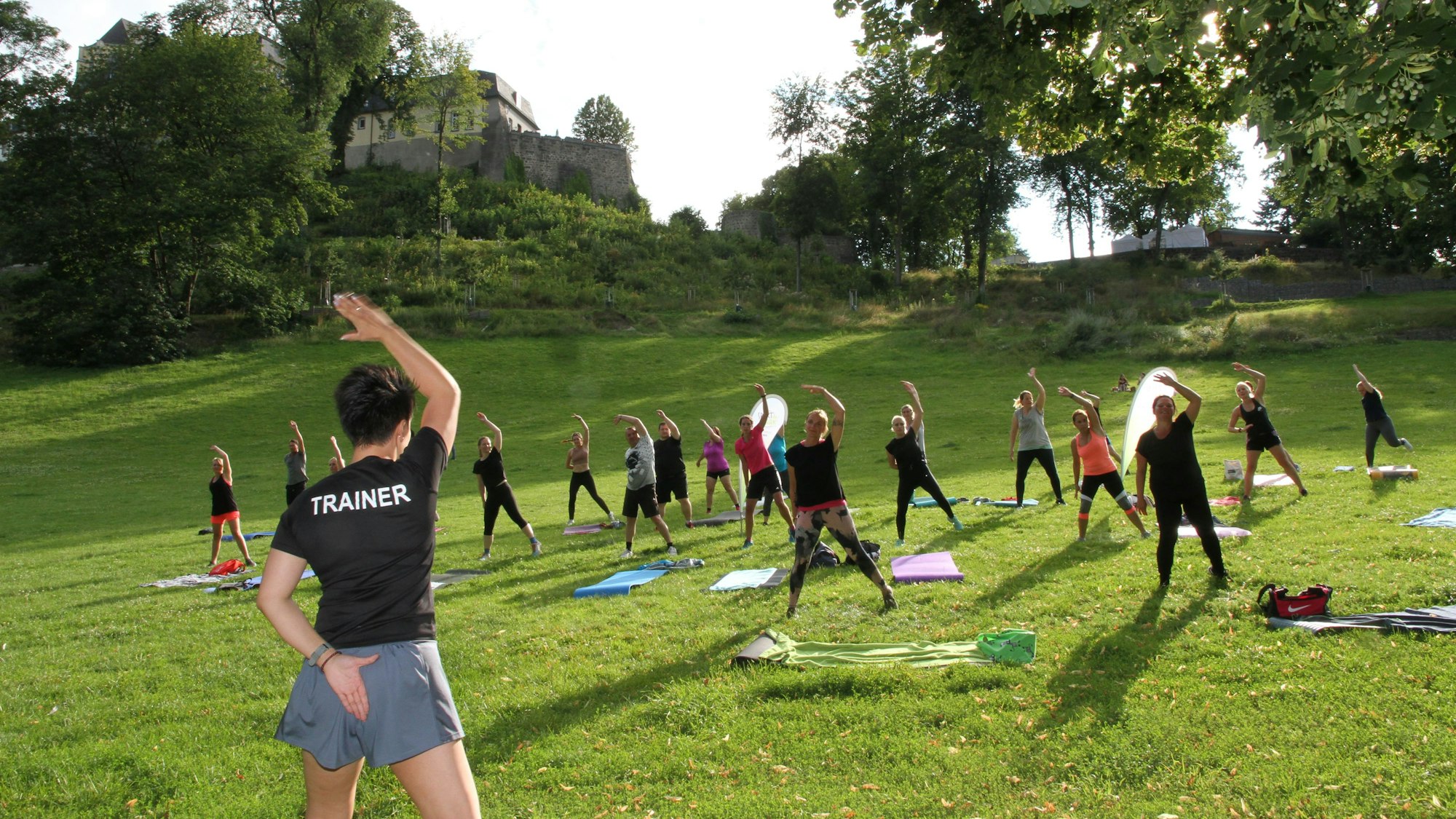 Menschen turnen auf einer Hangwiese. Eine Frau leitet das Training an, auf ihrem T-Shirt steht Trainer.