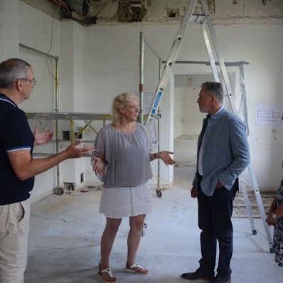 Zwei Männer und zwei Frauen stehen in einem Rohbau; am rechten Bildrand steht ein Transparent mit der Aufschrift "Förderverein Hebammenhaus Rhein-Sieg".