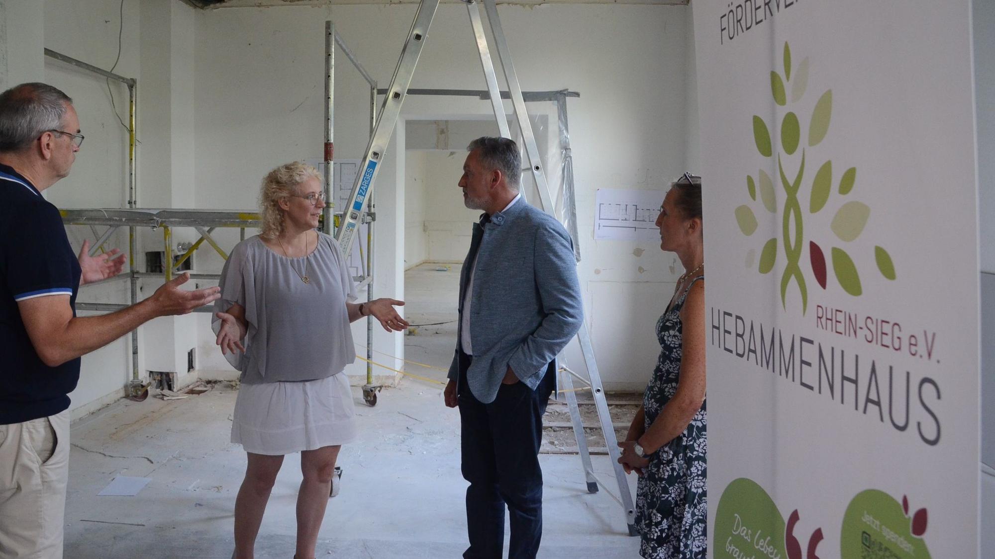 Zwei Männer und zwei Frauen stehen in einem Rohbau; am rechten Bildrand steht ein Transparent mit der Aufschrift "Förderverein Hebammenhaus Rhein-Sieg".