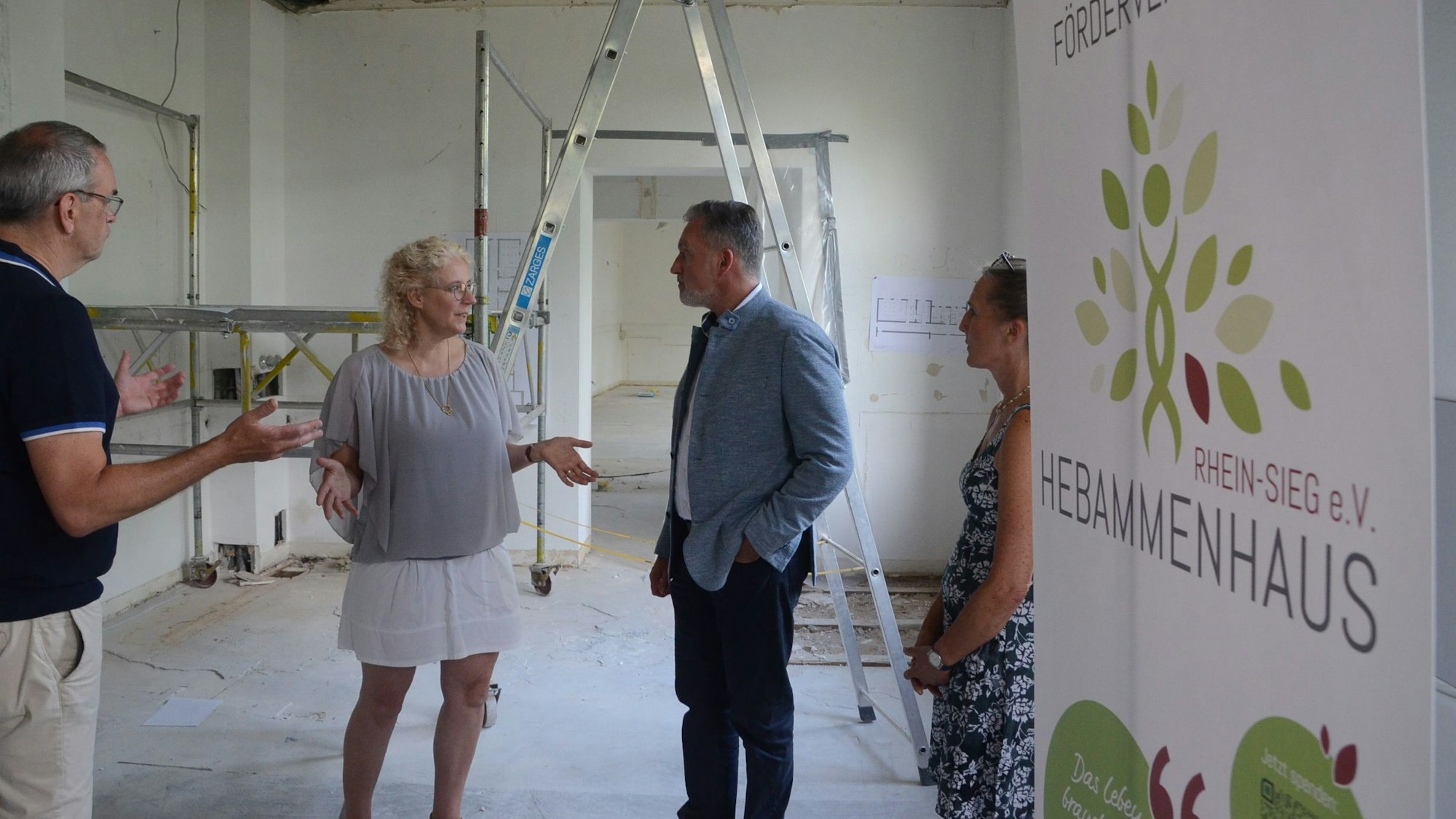 Zwei Männer und zwei Frauen stehen in einem Rohbau; am rechten Bildrand steht ein Transparent mit der Aufschrift "Förderverein Hebammenhaus Rhein-Sieg".