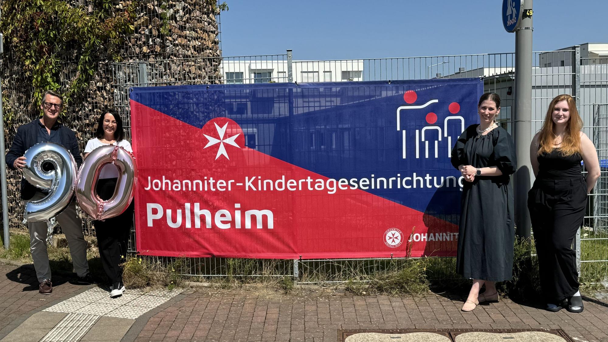 Vier Personen, zwei halten die Zahlen 9 und 0 in ihren Händen, zwischen ihnen ein Transparten mit der Aufschrift „Johanniter-Kindertageseinrichtung Pulheim“.