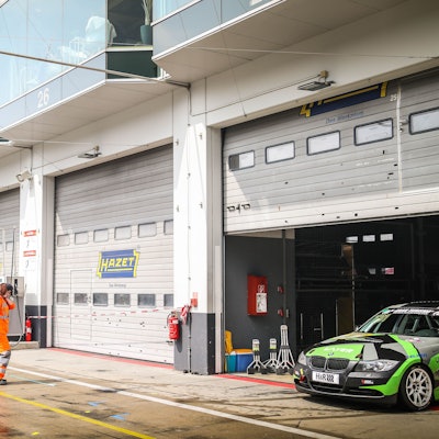 In der Boxengasse des Nürburgrings sind zwei Boxen mit rot-weißem Flatterband abgesperrt und geschlossen. Daneben sind Boxen geöffnet, in der rechten steht ein grün-schwarzer Rennwagen.