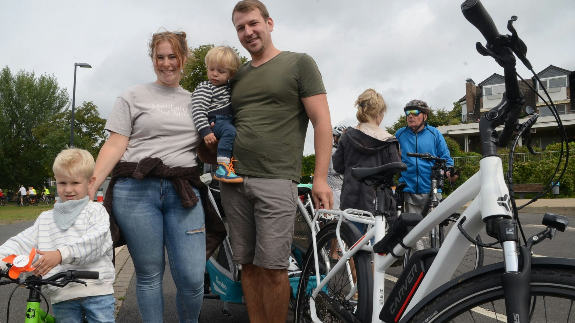 Zwei Erwachsene und zwei Kinder mit Fahrrädern. Ein Kind hat ein eigenes Fahrrad, das zweite sitzt beim Vater auf dem Arm.