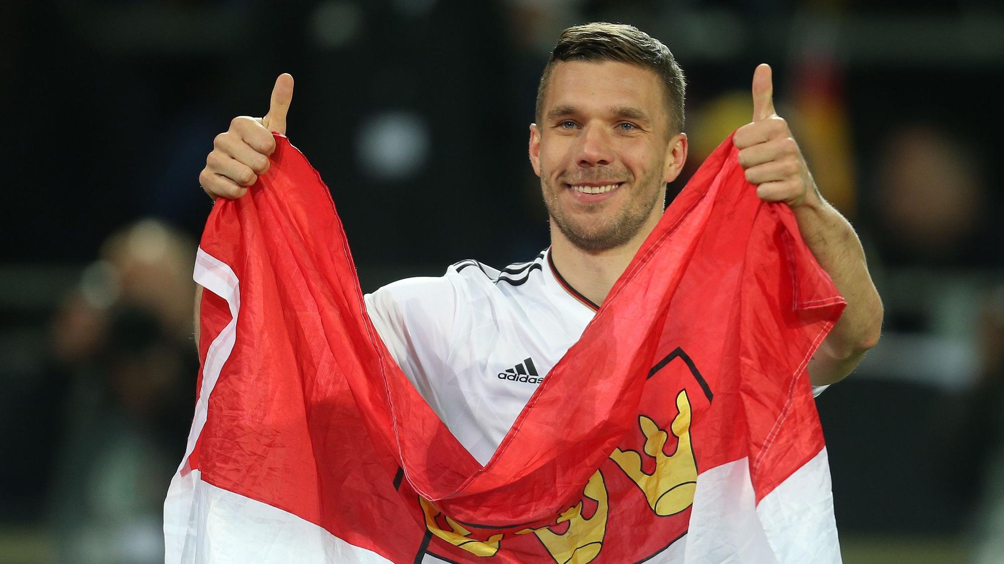 ukas Podolski verabschiedet sich nach dem Spiel von den Fans und hält dabei eine Kölner Fahne.