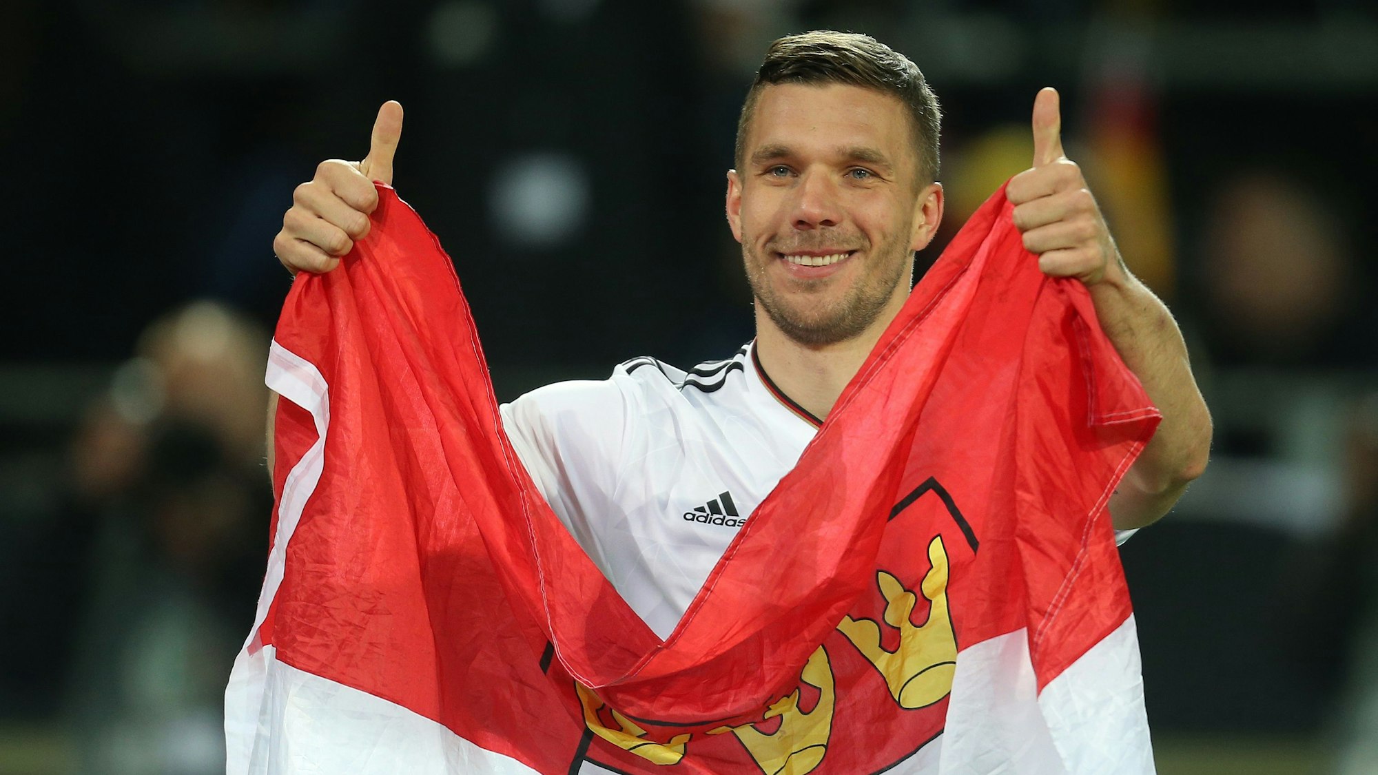 ukas Podolski verabschiedet sich nach dem Spiel von den Fans und hält dabei eine Kölner Fahne.