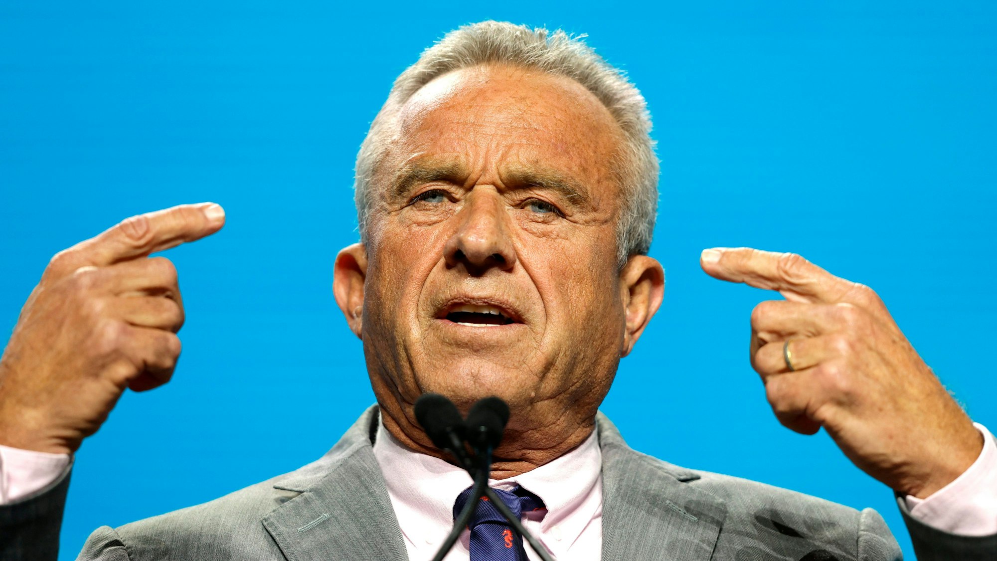 Robert F. Kennedy Jr. steht vor einem blauen Hintergrund und gestikuliert während einer Rede.